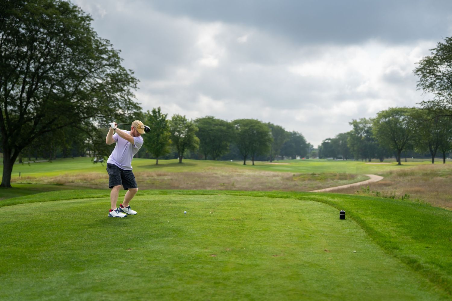 Golfer swinging a club on a lush green golf course under a cloudy sky surrounded by trees.