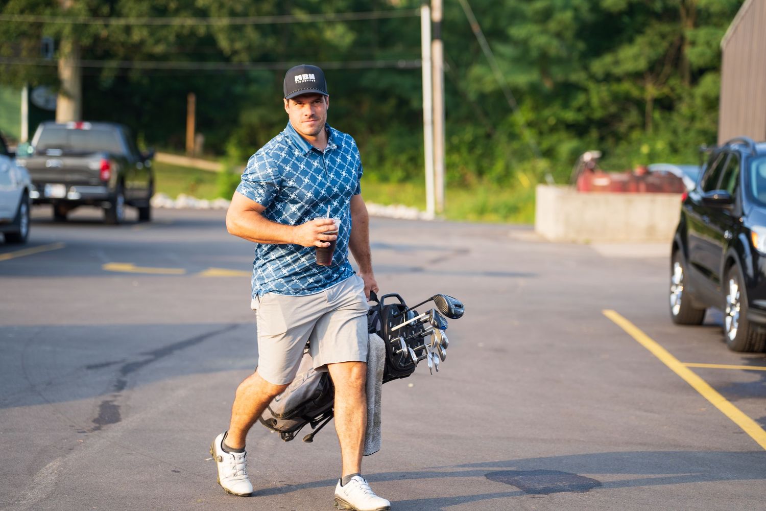 Man in blue polo shirt carrying golf bag across parking lot on sunny day.