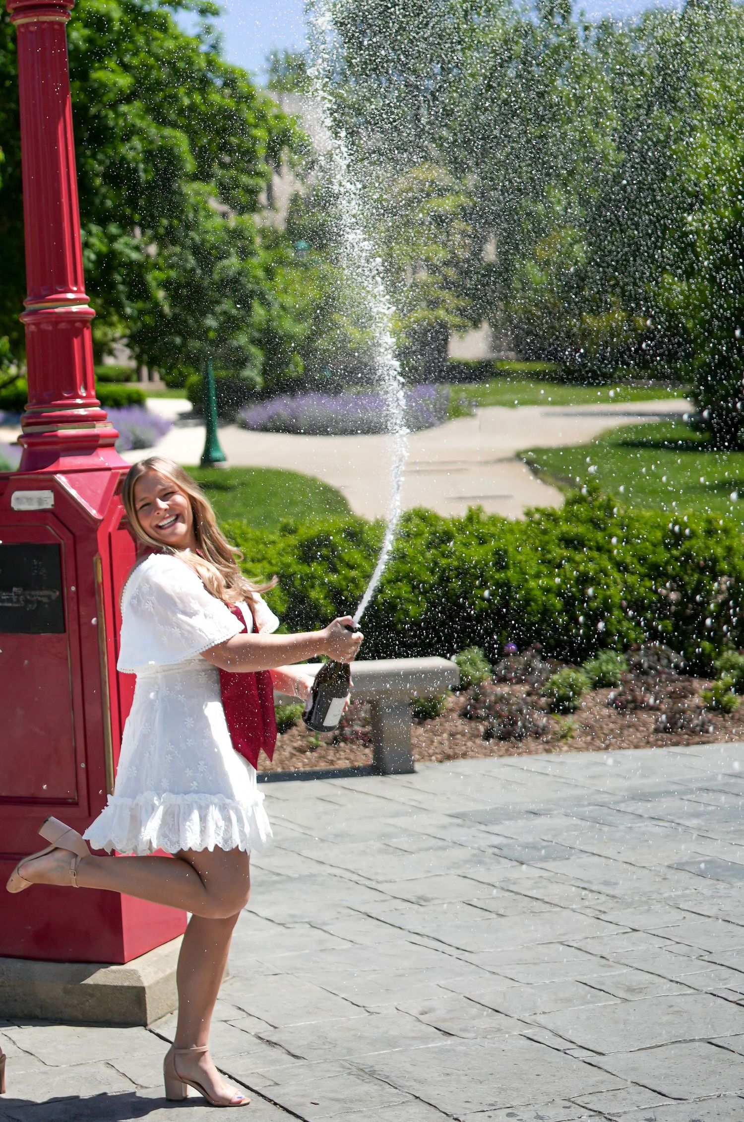 Joyful blonde woman in white dress popping champagne bottle near red lamppost on sunny campus.