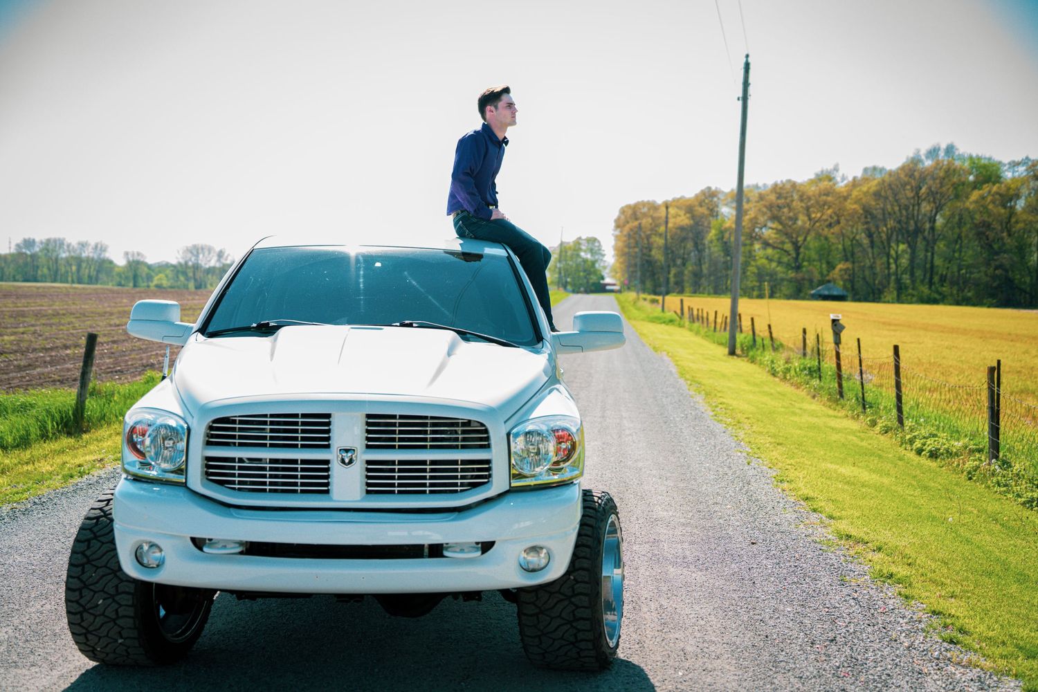Young man sitting on hood of lifted white Dodge Ram truck on a scenic rural gravel road.