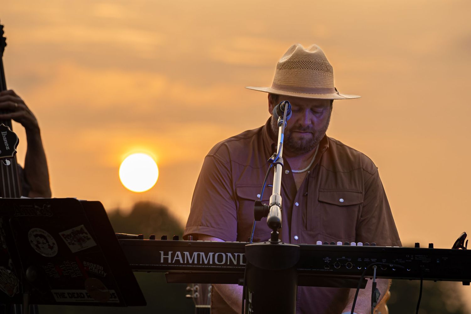 Bearded musician playing Hammond organ at golden hour outdoor concert wearing wide-brim hat.