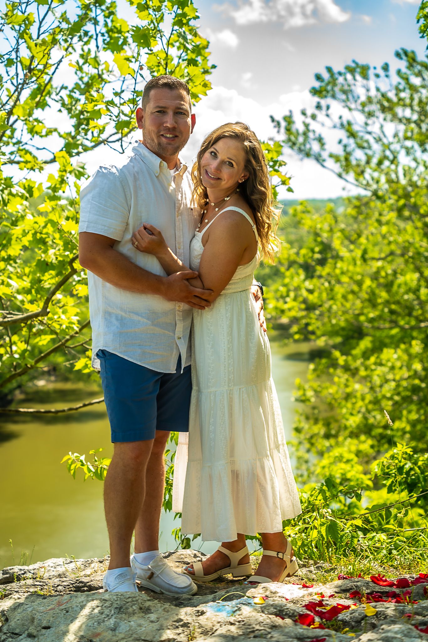 Happy couple posing on rocky ledge with river and green trees in sunny background.
