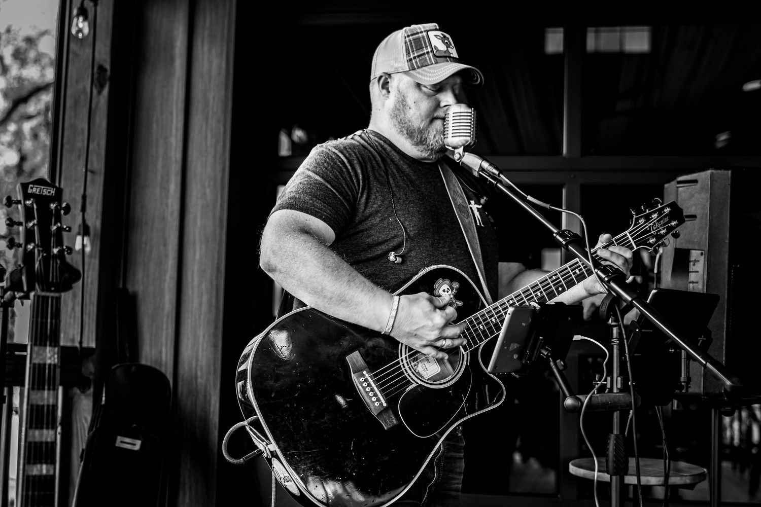 Black and white photo of heavyset guitarist in cap singing into microphone while playing black acoustic guitar.