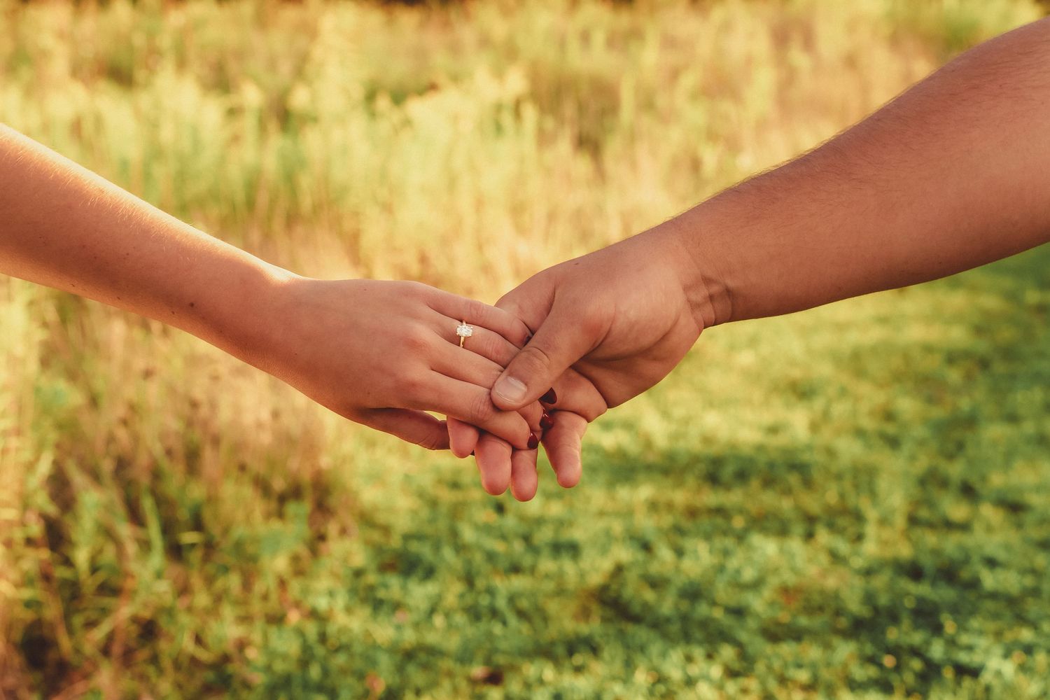 Couple holding hands in a sunlit grassy field, showcasing an engagement ring.