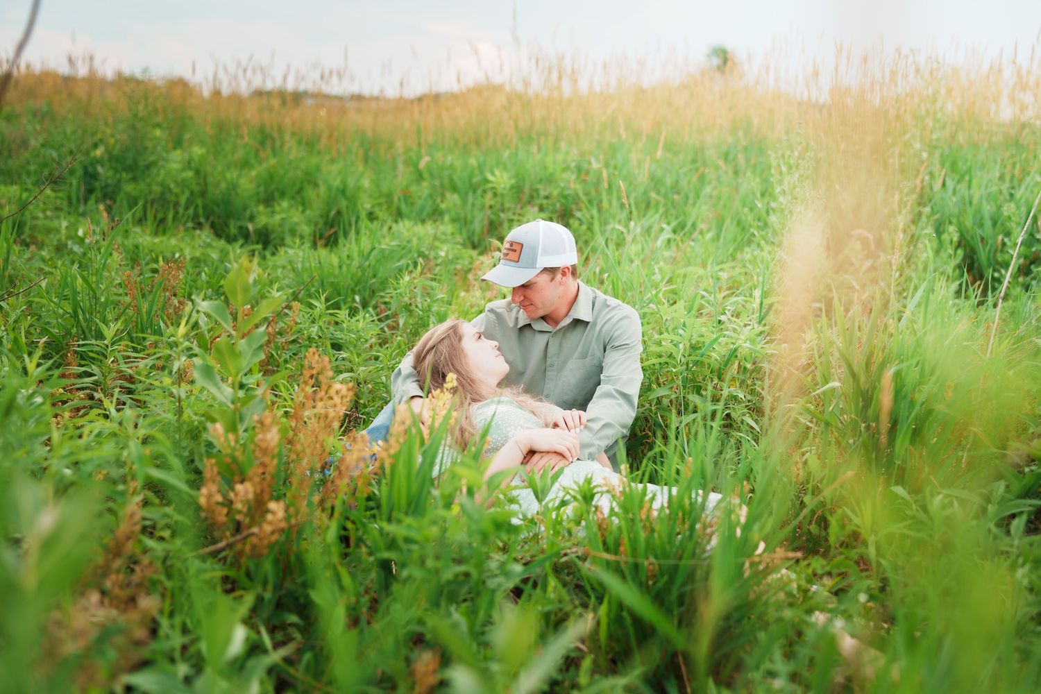 Loving couple sitting together in tall green grass during golden hour sunset.