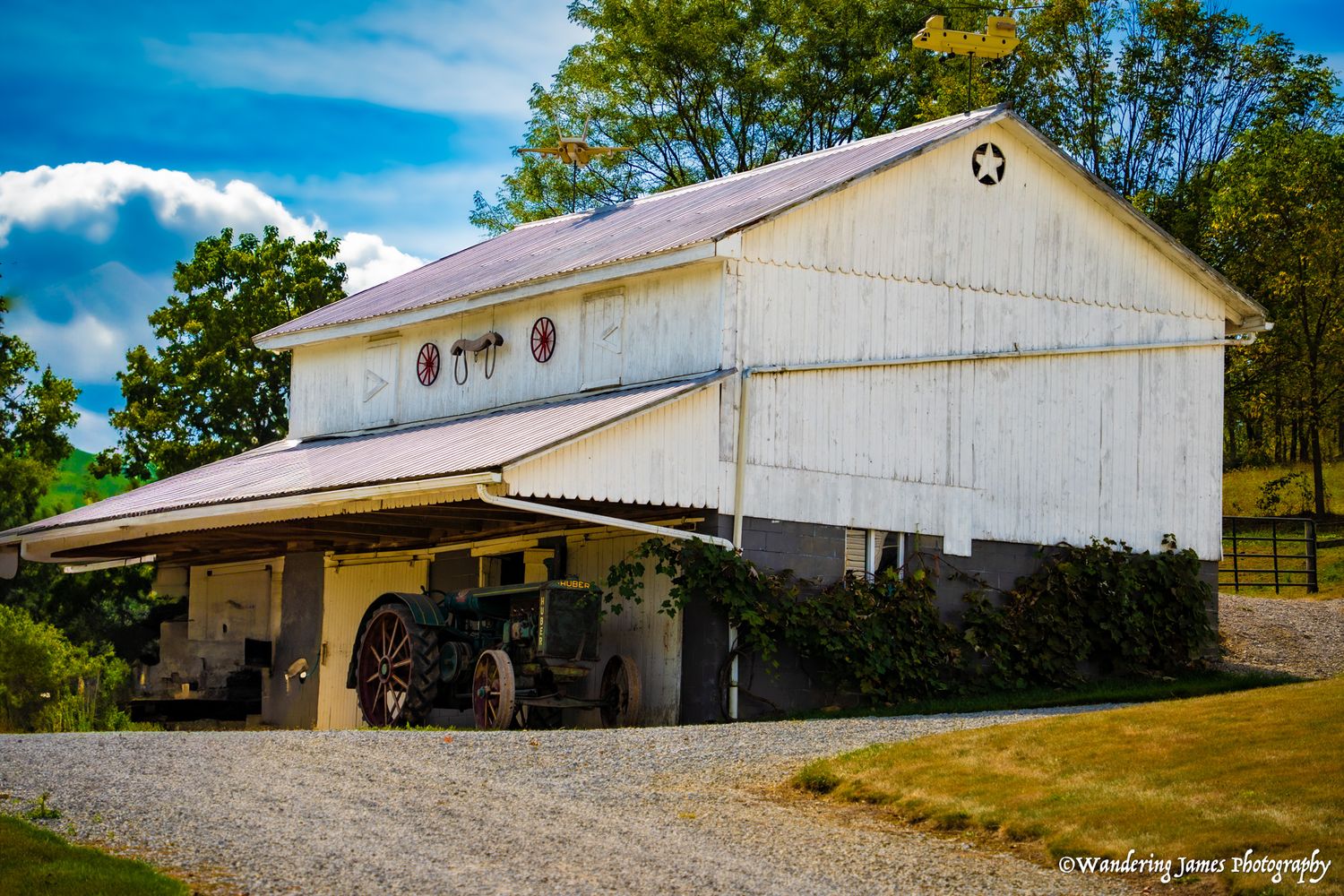 A vintage white barn with old farm equipment sits under a blue sky.