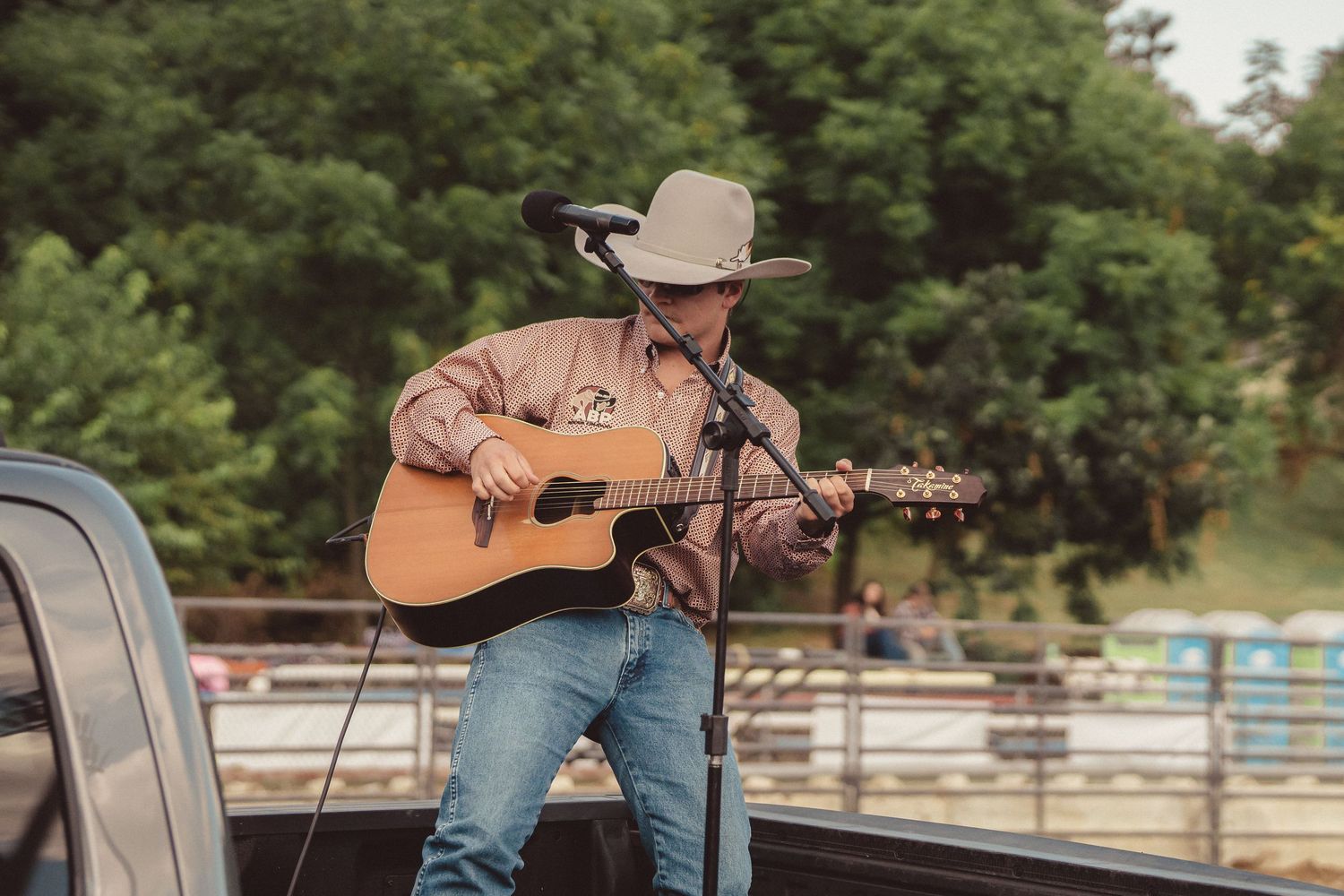 Country musician in cowboy hat performing acoustic guitar on truck bed at outdoor rodeo.
