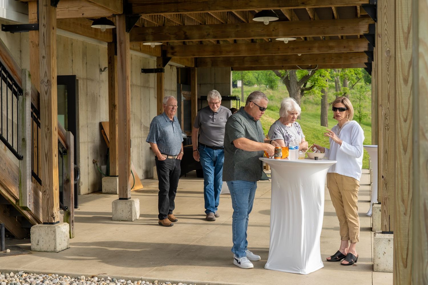Small group of people socializing around a tall cocktail table on a rustic wooden covered porch at an outdoor event.
