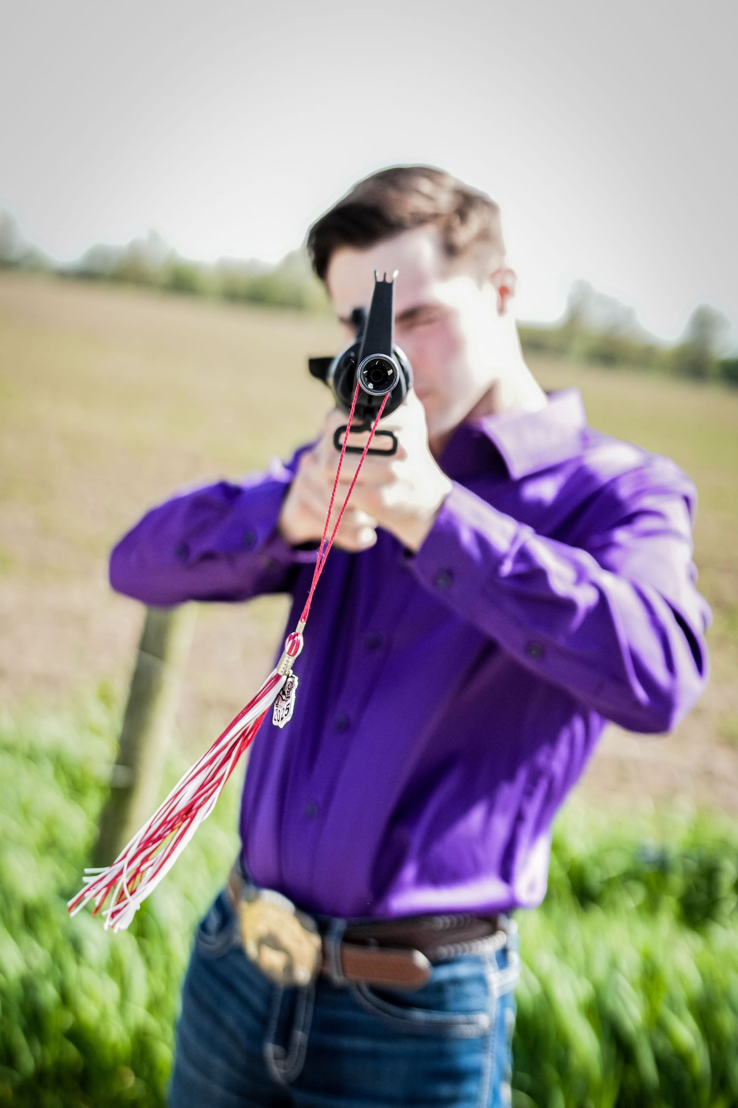 Young man in purple shirt aims a starting pistol with red tassel toward camera in a field.
