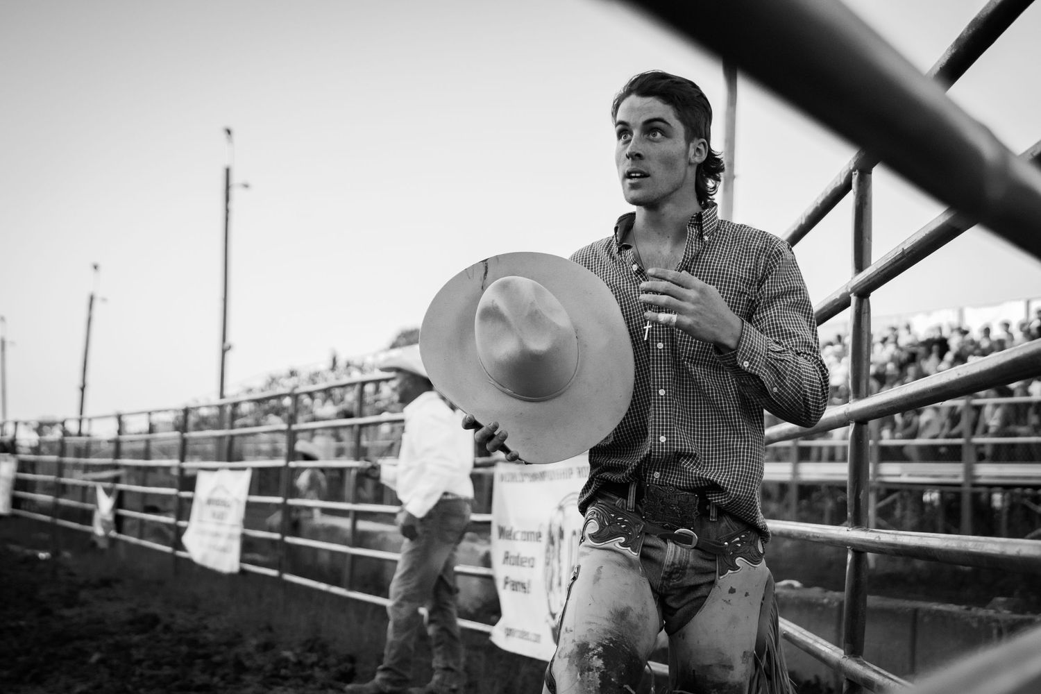 Black and white photo of a cowboy holding a hat at a rodeo arena with crowded bleachers in the background.