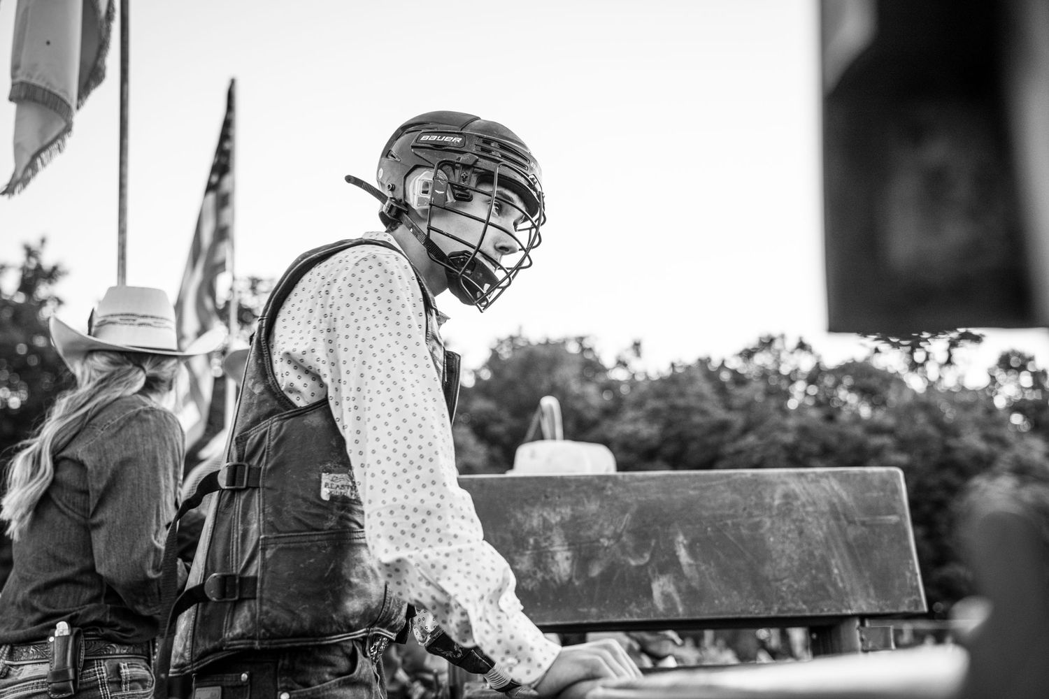 A black and white portrait of a rodeo bull rider wearing a helmet and protective vest behind the chutes.