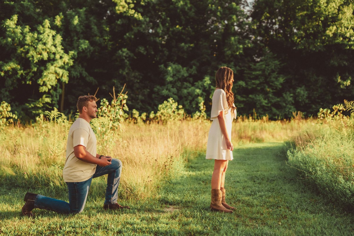 Man kneeling on one knee proposing to woman in white dress on scenic green meadow path surrounded by trees.
