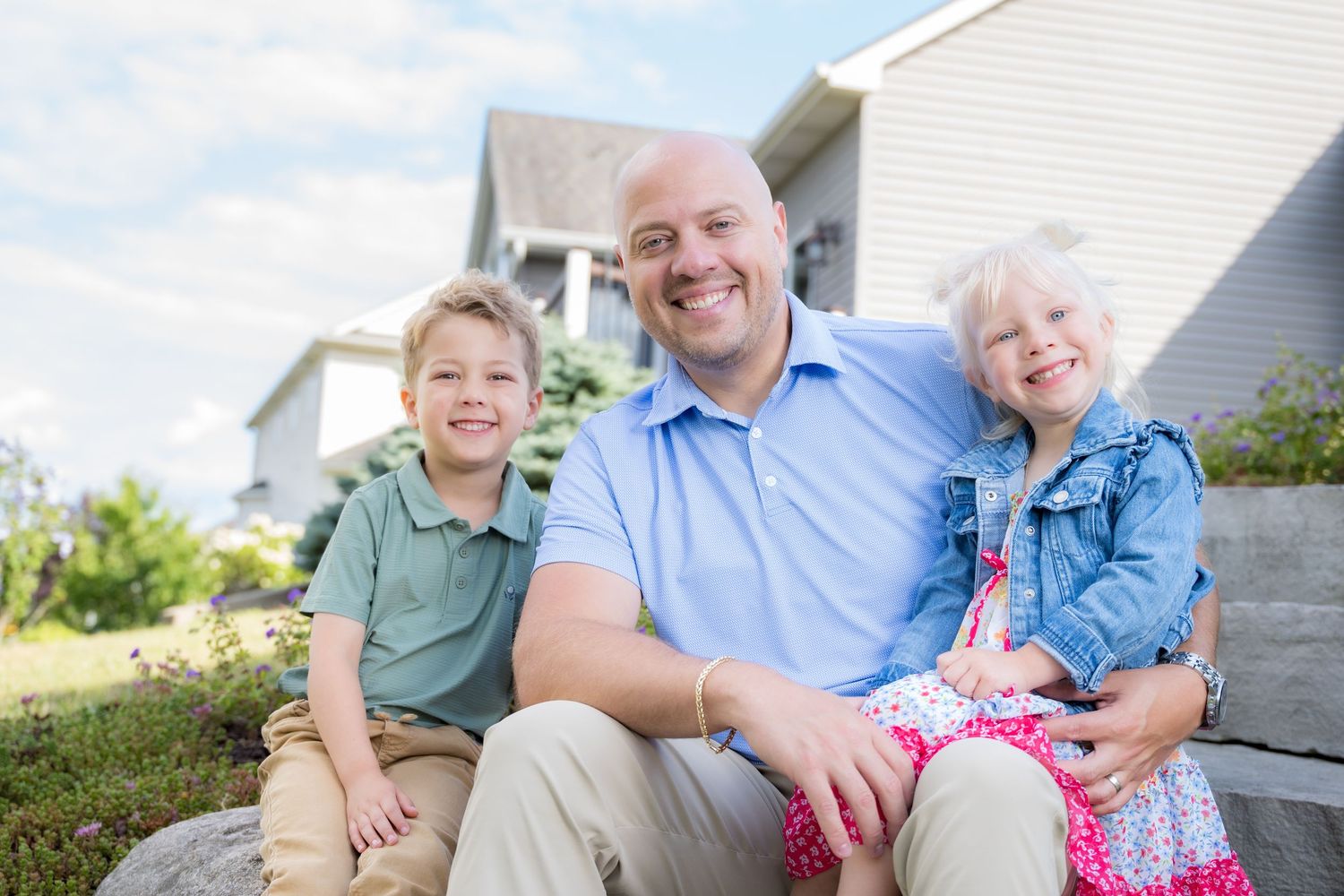 A smiling father sits outdoors with his two young children, a boy and girl, in front of a suburban home.