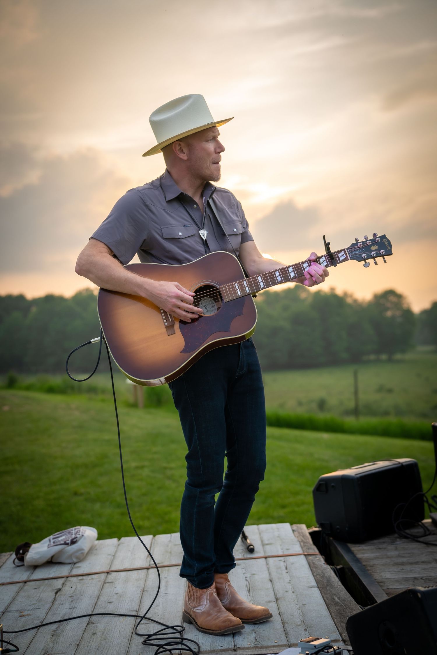 A country musician in a white hat plays acoustic guitar on an outdoor stage at sunset.
