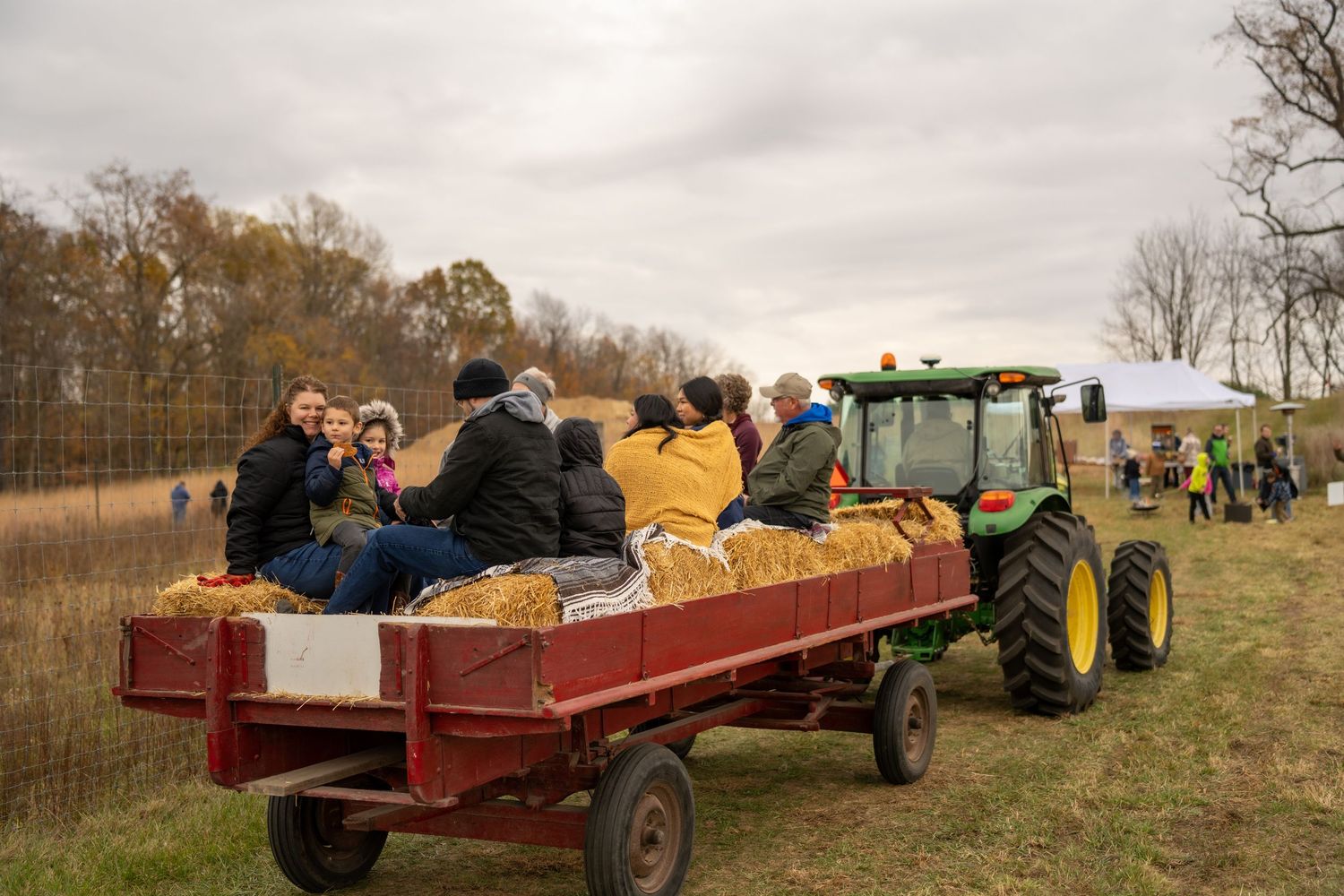 Families enjoy a hay ride on a red wagon pulled by a green John Deere tractor through an autumn field.