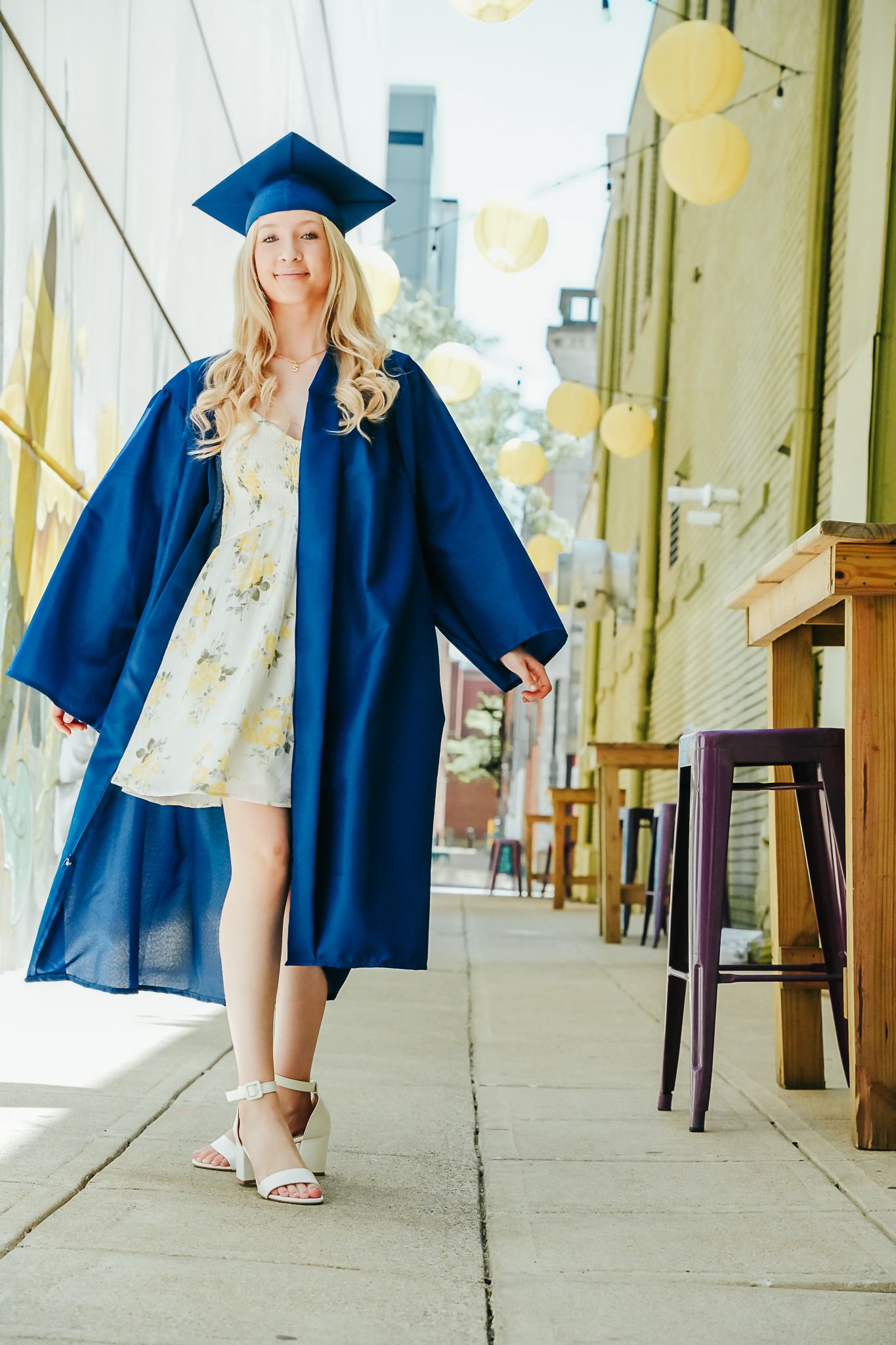 Blonde graduate in blue cap and gown walking along sunny outdoor pathway in floral dress.