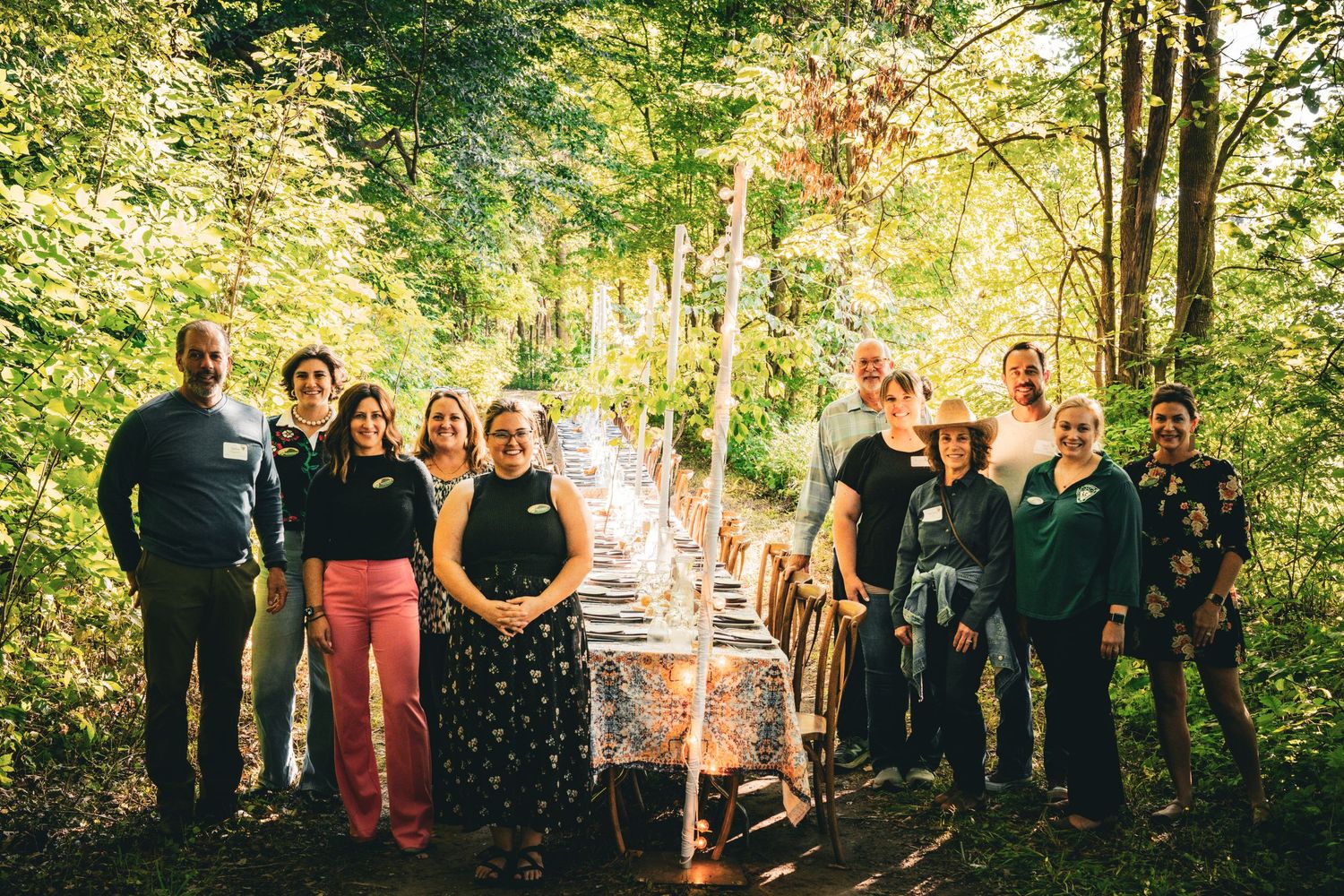 Group of smiling guests posing beside a long decorated outdoor dining table set in a sunlit forest clearing.