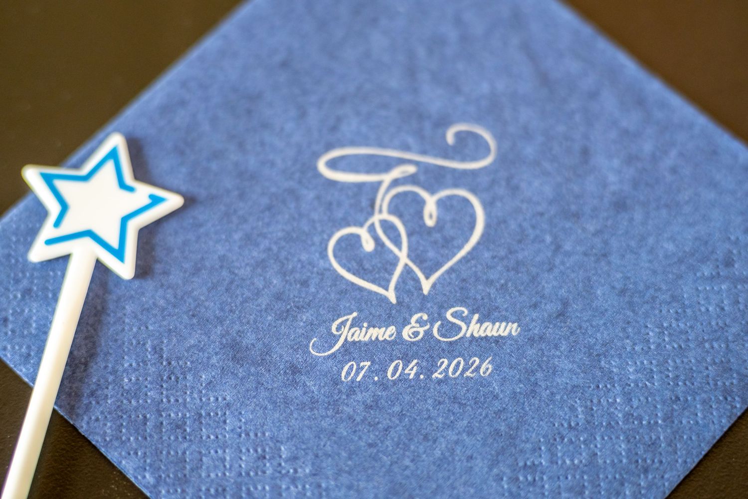 Blue personalized wedding napkin with intertwined hearts, names Jaime & Shawn, date 07.04.2026, beside a star wand.