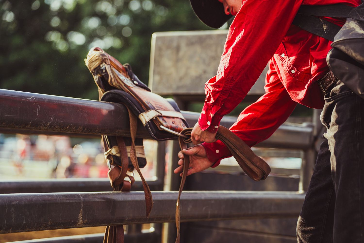 Cowboy in red shirt adjusting rodeo bull riding equipment on fence at outdoor western event.