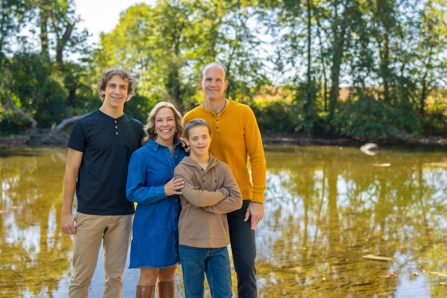 Family of four smiling outdoors near a reflective pond surrounded by lush green trees on a sunny day.