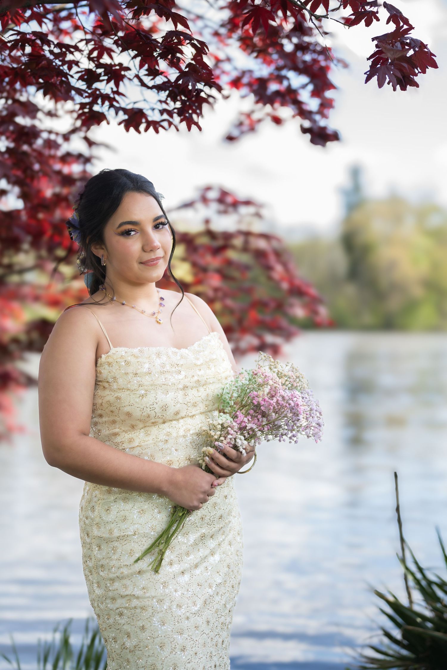 Young woman in cream floral dress holding bouquet near lake under red maple tree.