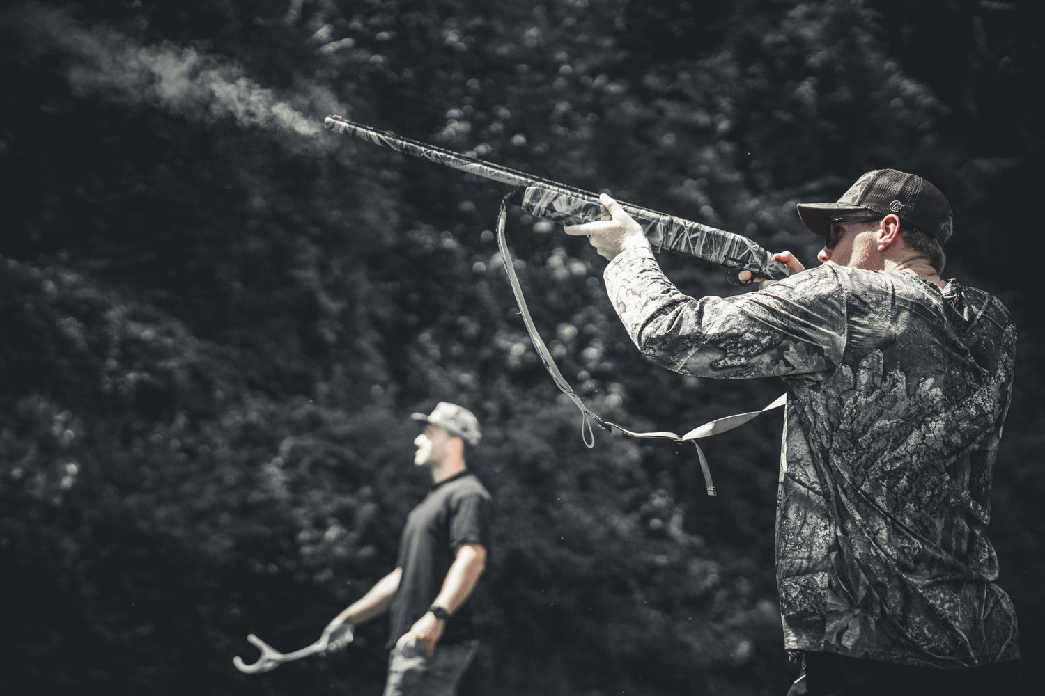 Two hunters in camouflage clothing shoot shotguns outdoors, with smoke visible from the barrel during a hunting session.