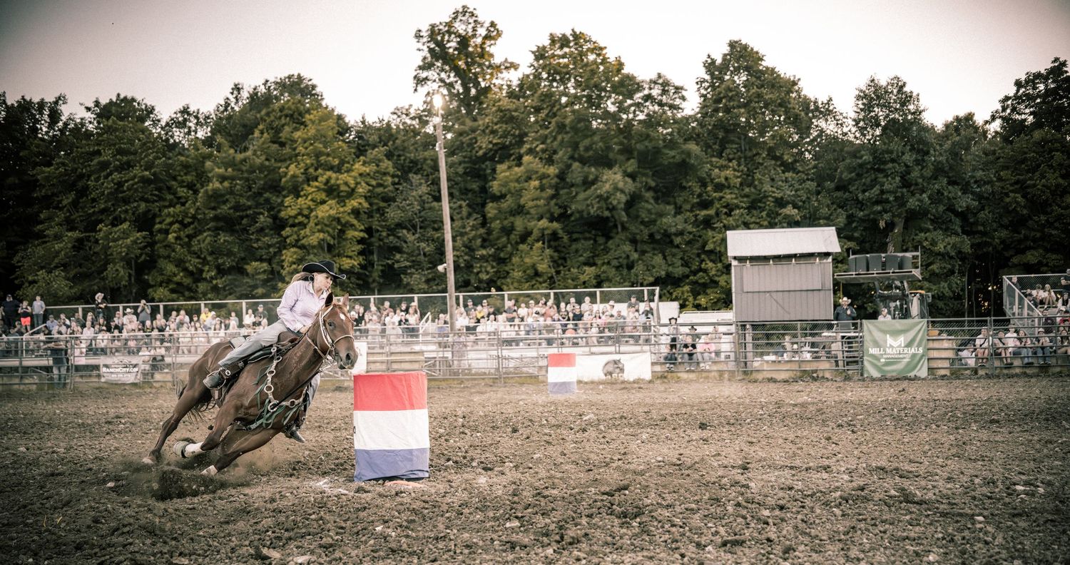 Barrel racer on horseback navigates around a red and blue barrel at a dusty outdoor rodeo event.