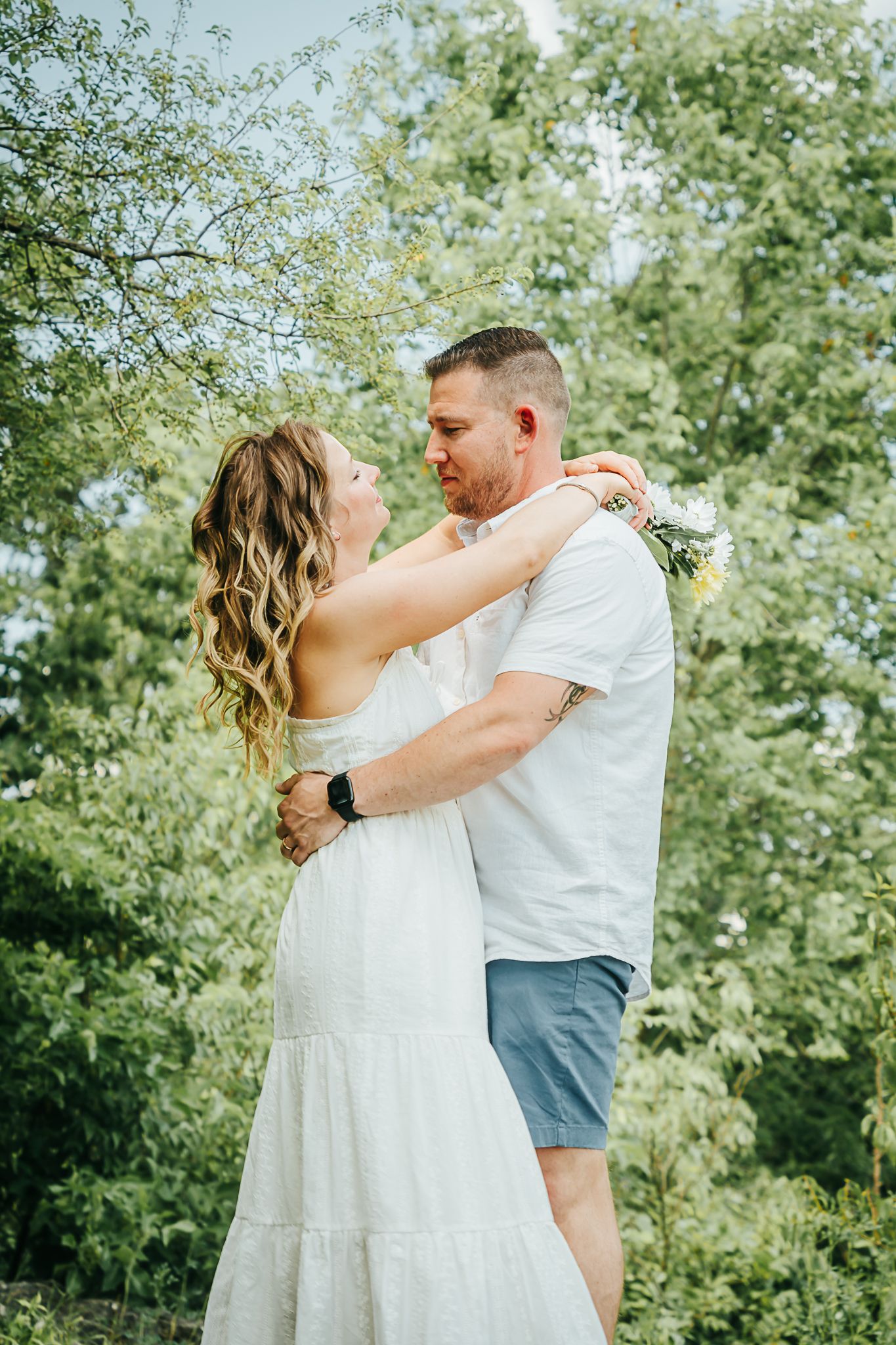 Couple holding each other close outdoors surrounded by lush green trees on a sunny day.
