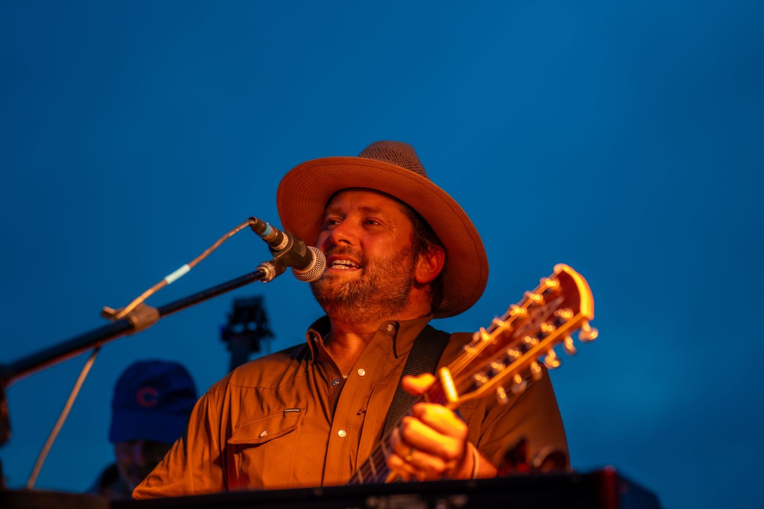 Male vocalist singing into microphone playing instrument at outdoor night concert in wide hat.