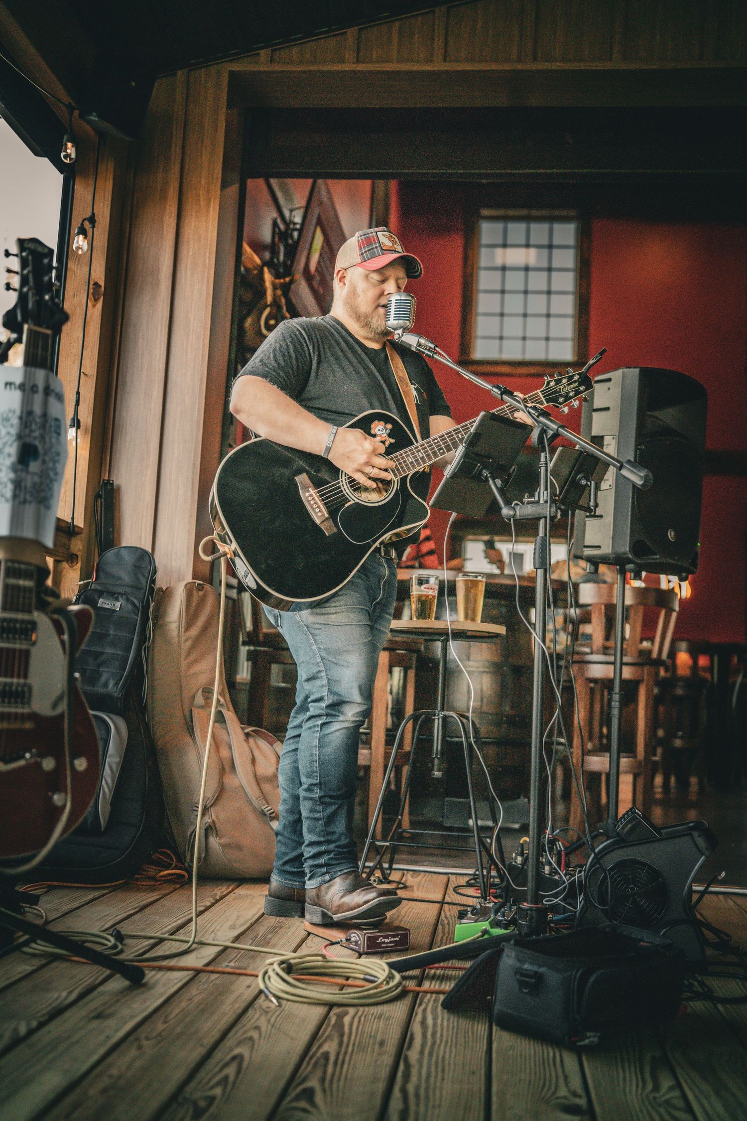 Heavyset guitarist in trucker cap plays black acoustic guitar on rustic wooden porch stage with multiple guitars nearby.