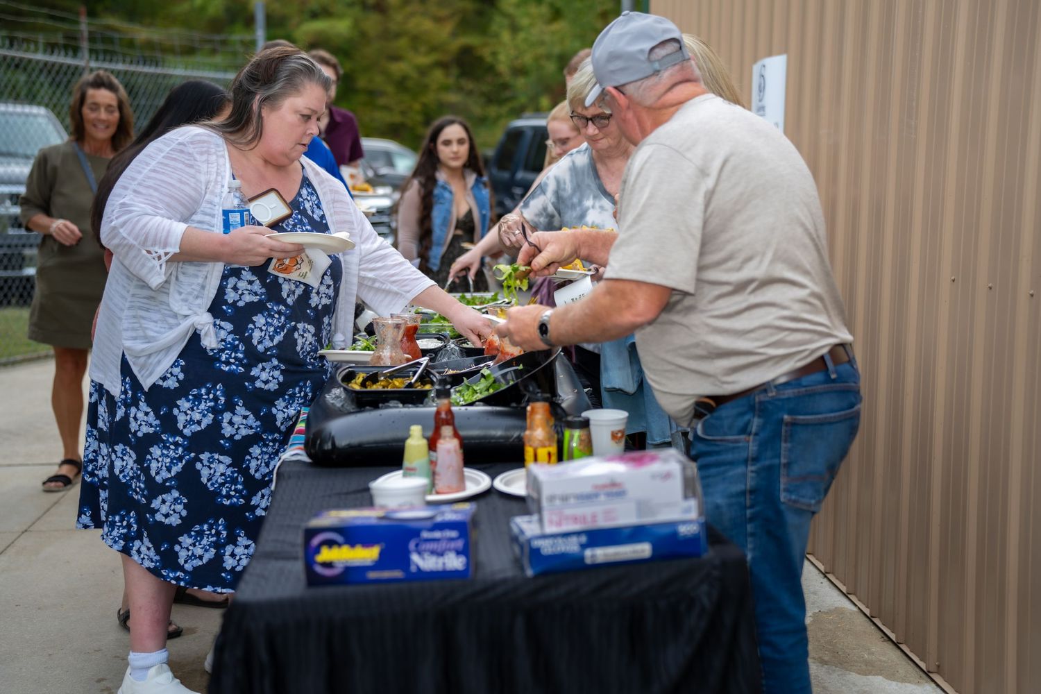 Man and woman serve themselves food at an outdoor buffet table during a casual community gathering event.