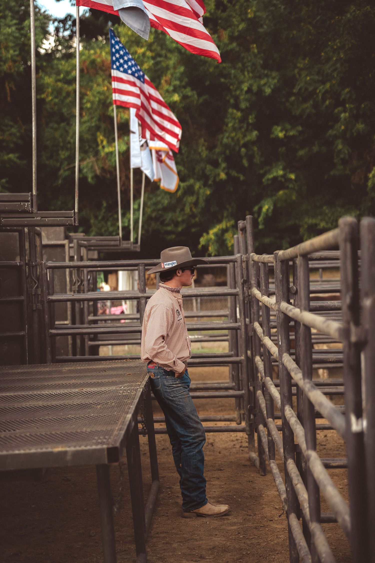 Young cowboy in hat and pink shirt leaning on metal fence at rodeo with American flags above.