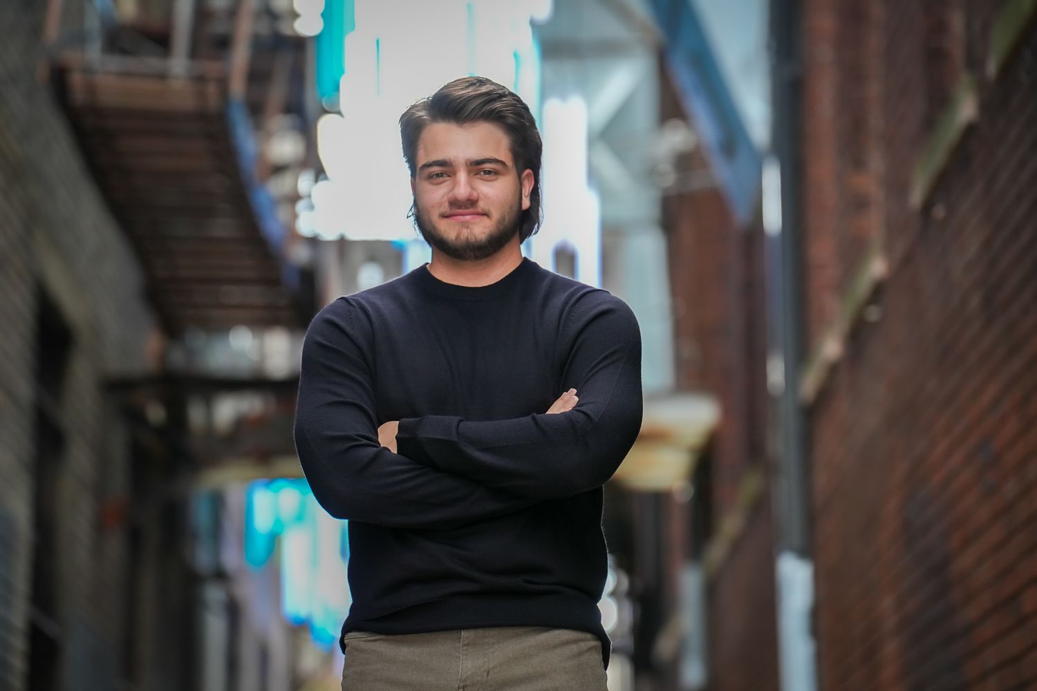 Young man with beard in black sweater standing with arms crossed in urban brick alleyway.