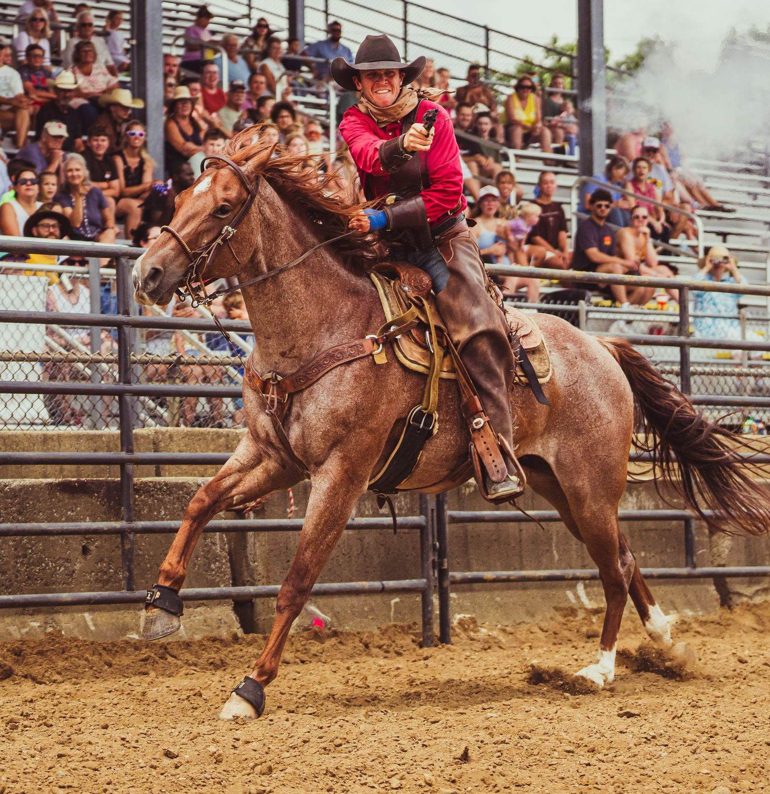 Cowboy in red shirt rides brown horse at full gallop before packed rodeo stadium crowd.