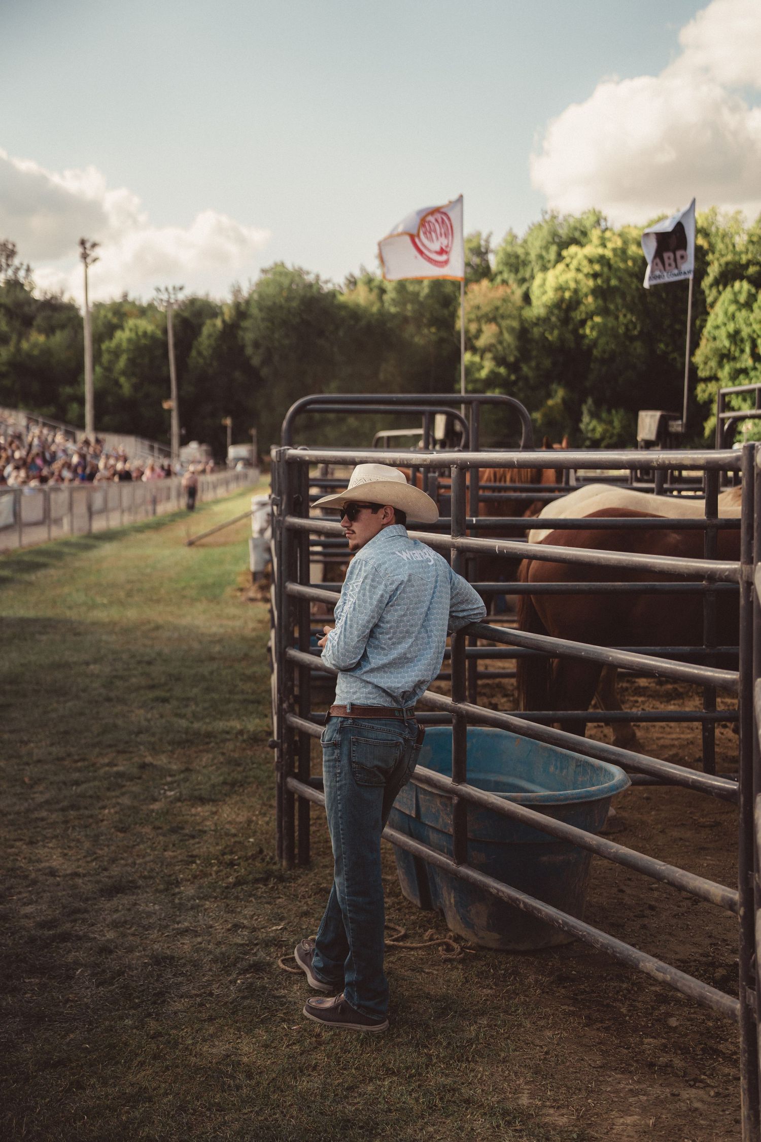 Cowboy in white hat and jeans leans on metal rodeo pen fence beside a horse at a western rodeo event.