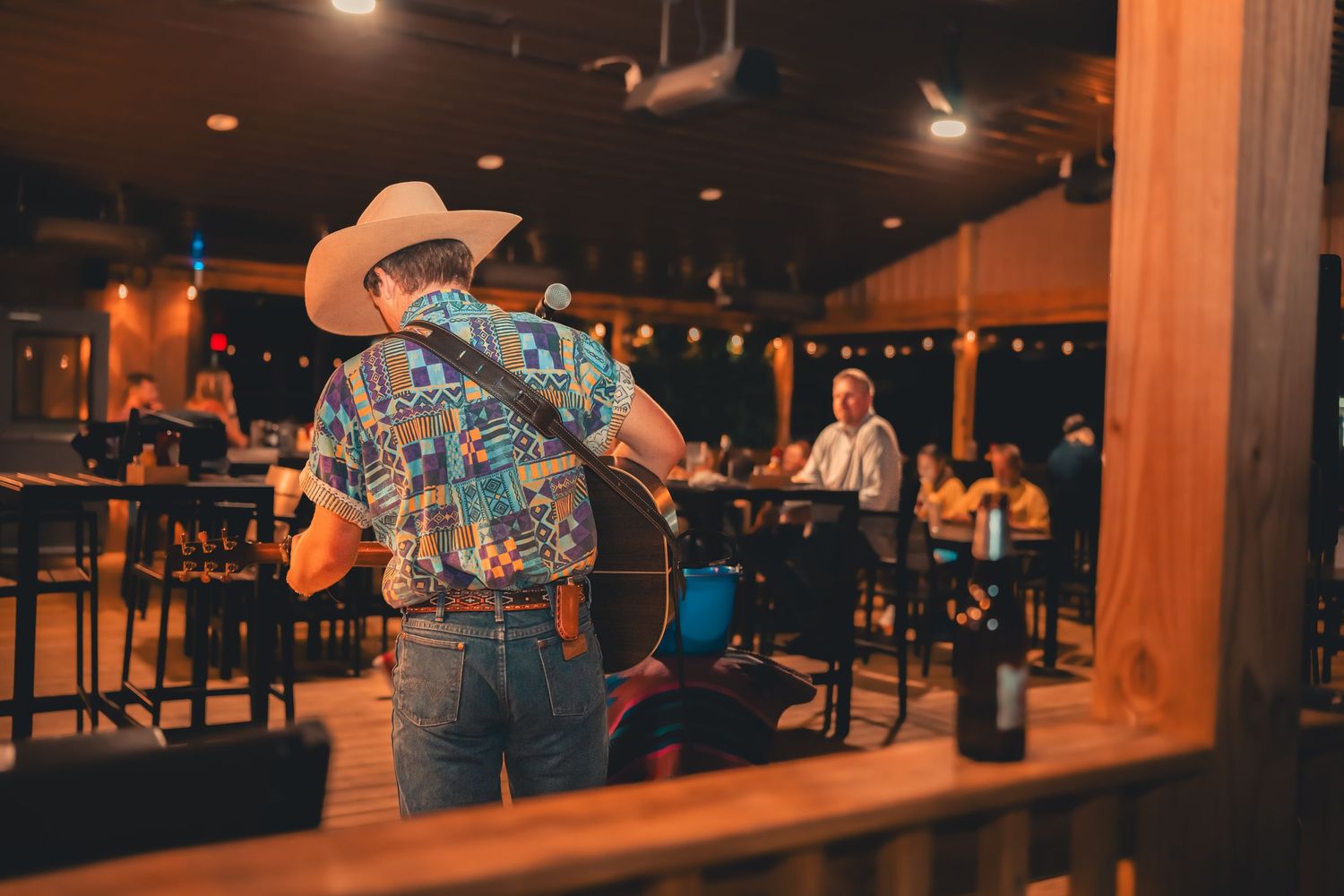 Country musician in cowboy hat and colorful shirt plays acoustic guitar on indoor stage for seated audience.