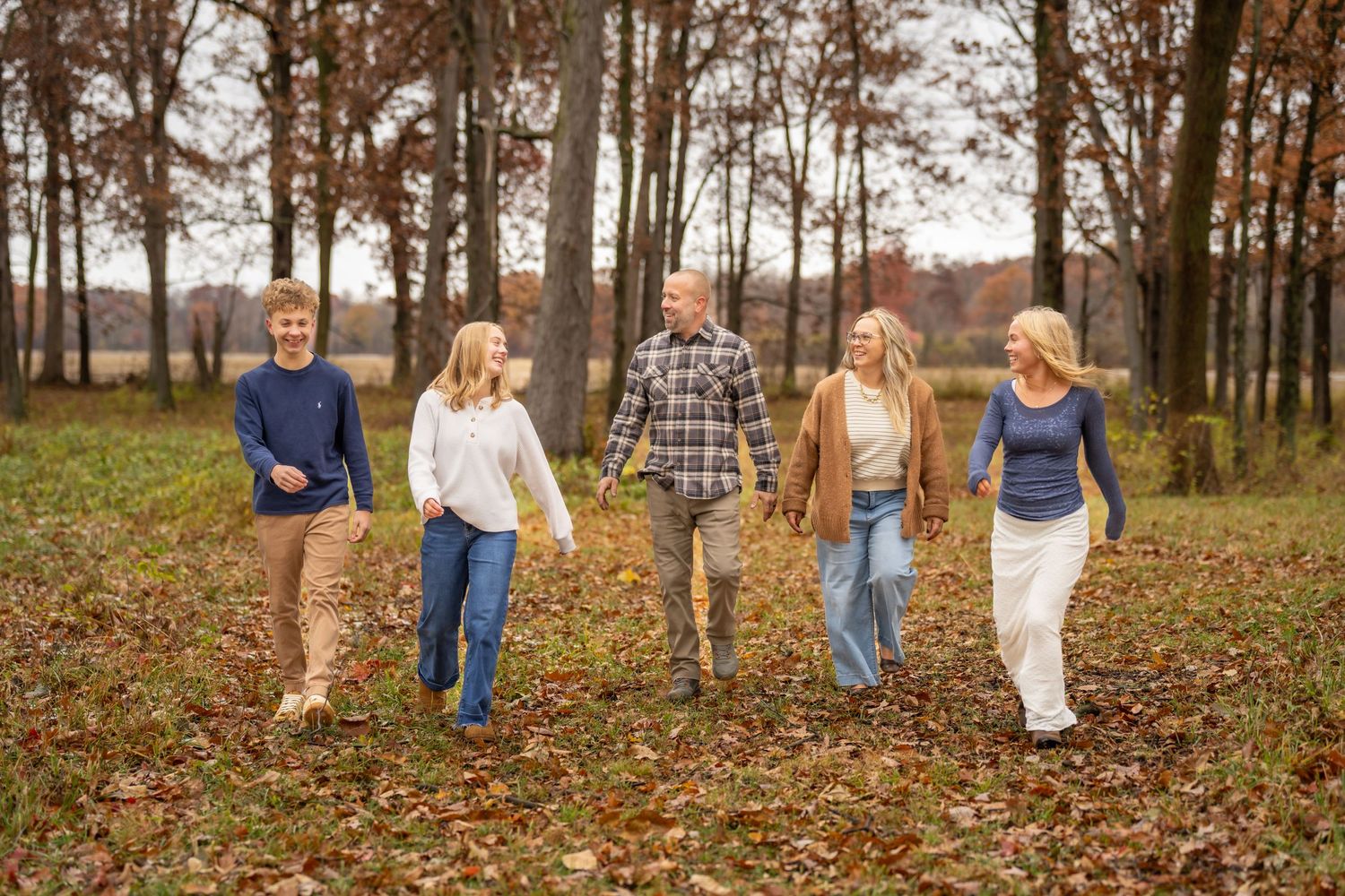 A family of five walks together through a leaf-covered field surrounded by tall autumn trees.
