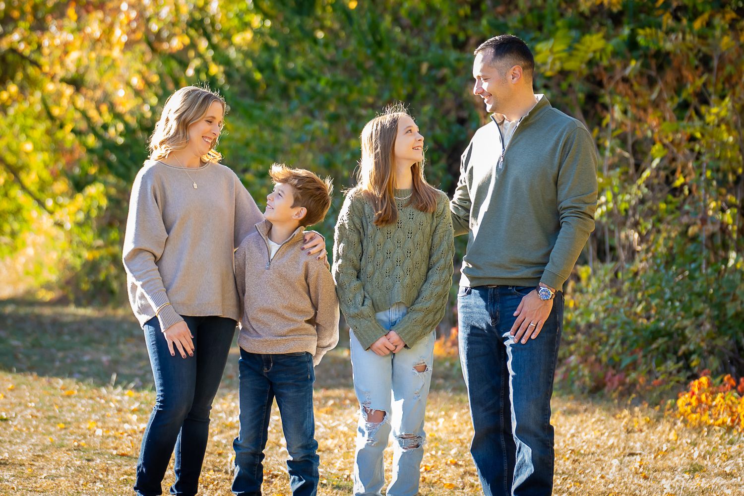 A family of four smiles at each other while standing outdoors in a sunny autumn setting.