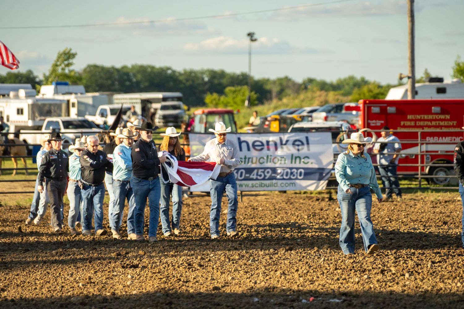 Group of cowboys carrying a banner for Henkel Construction Inc. across a sunny rodeo arena dirt floor.