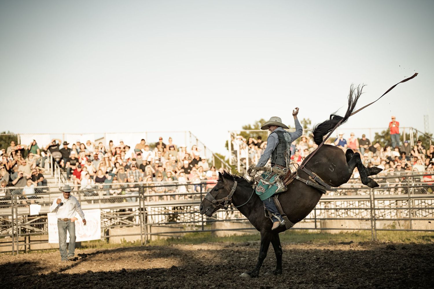 Rodeo cowboy in hat riding a bucking horse with one arm raised before a packed crowd of spectators.