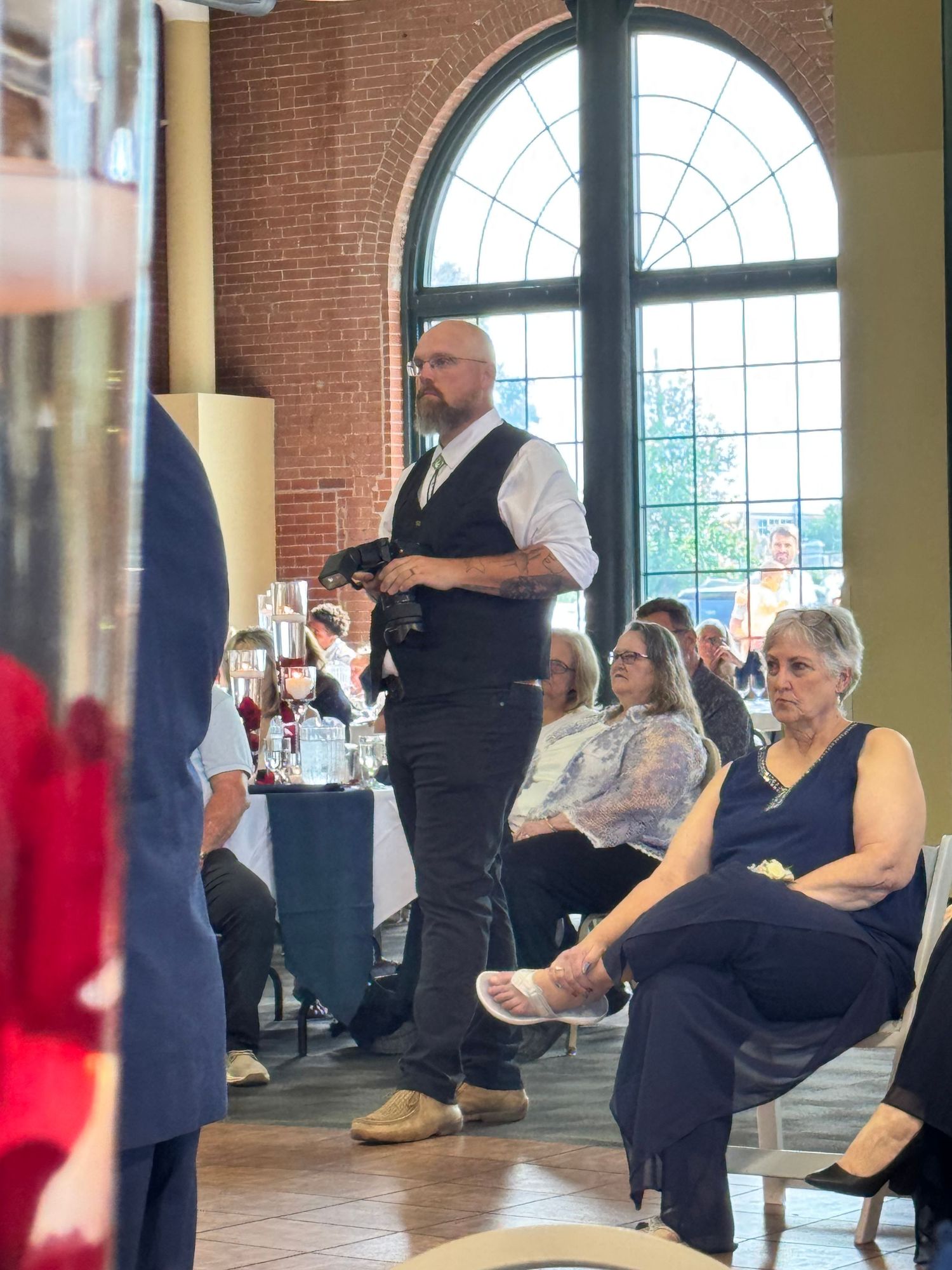 Bald man in vest stands at indoor event venue with seated guests and large arched windows in background.