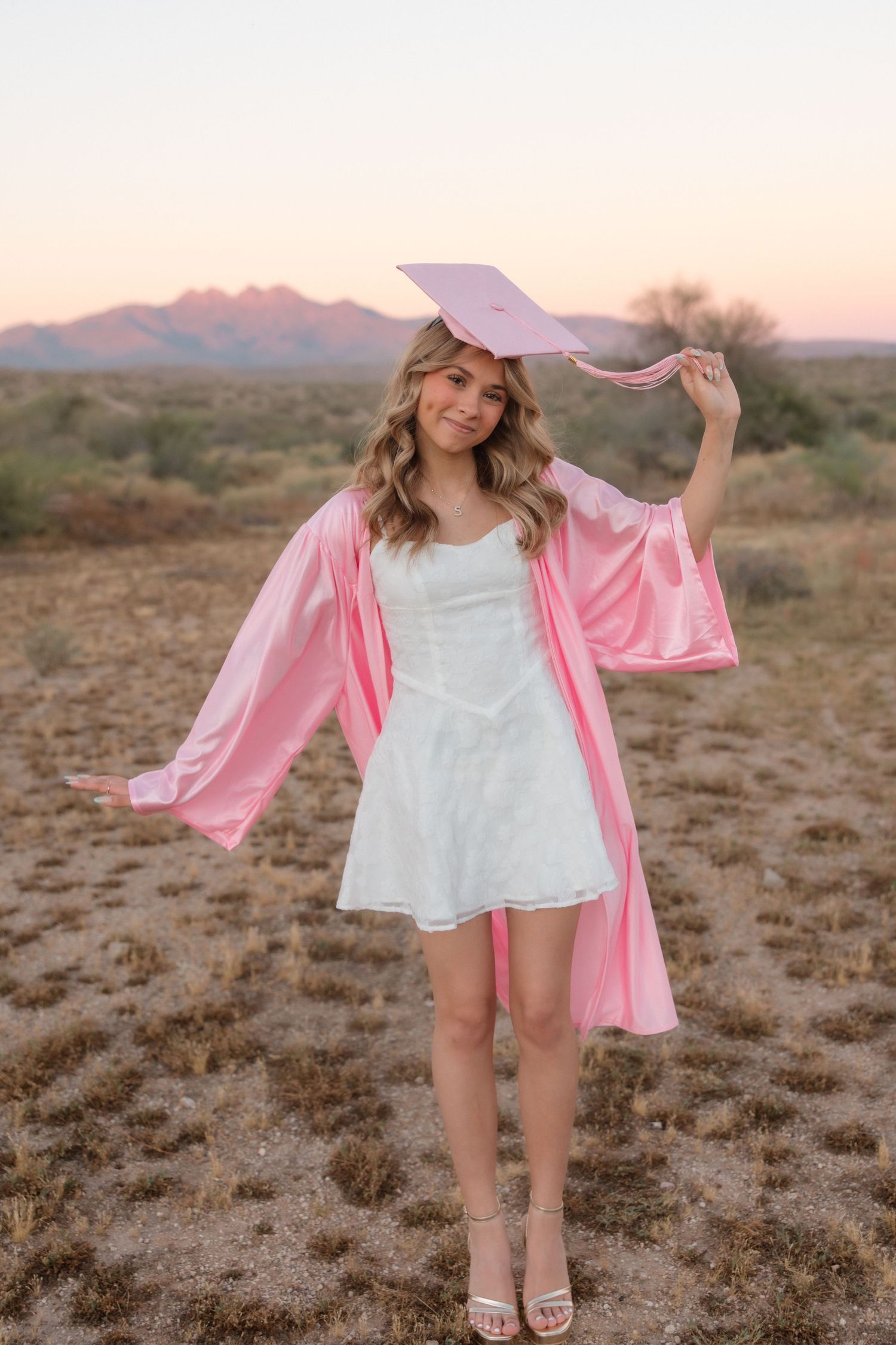 Young woman in pink graduation cap and gown over white dress poses in desert landscape at sunset.