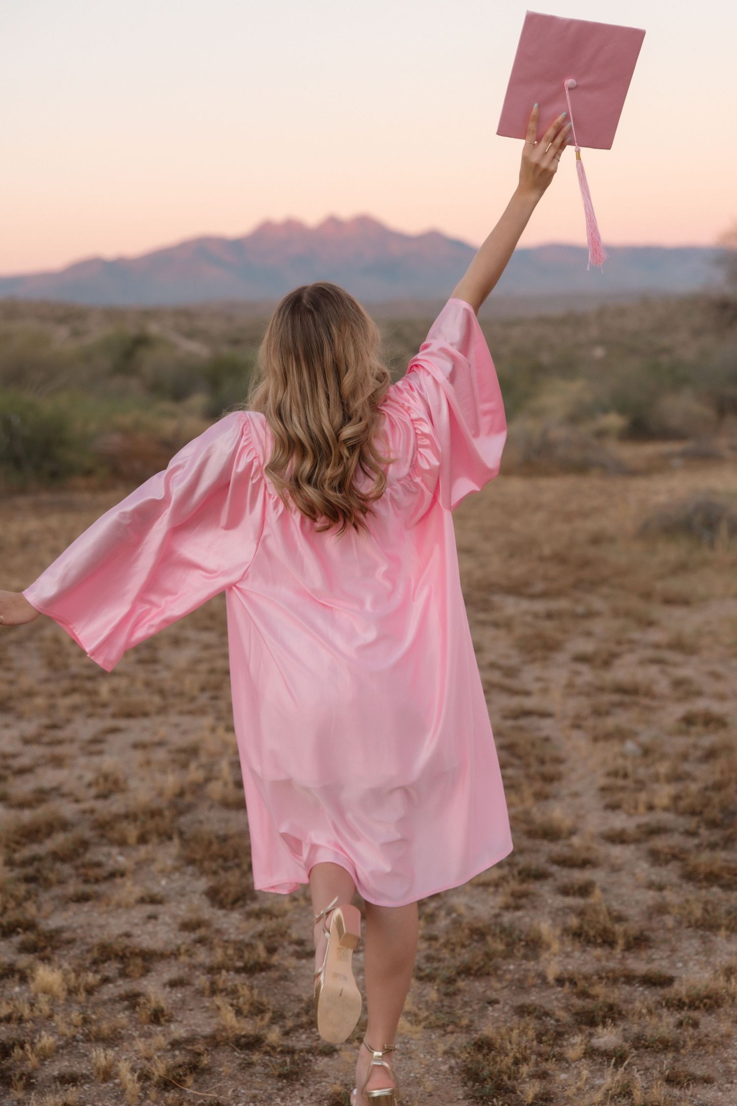 Graduate in pink cap and gown raises cap triumphantly in desert landscape at sunset.