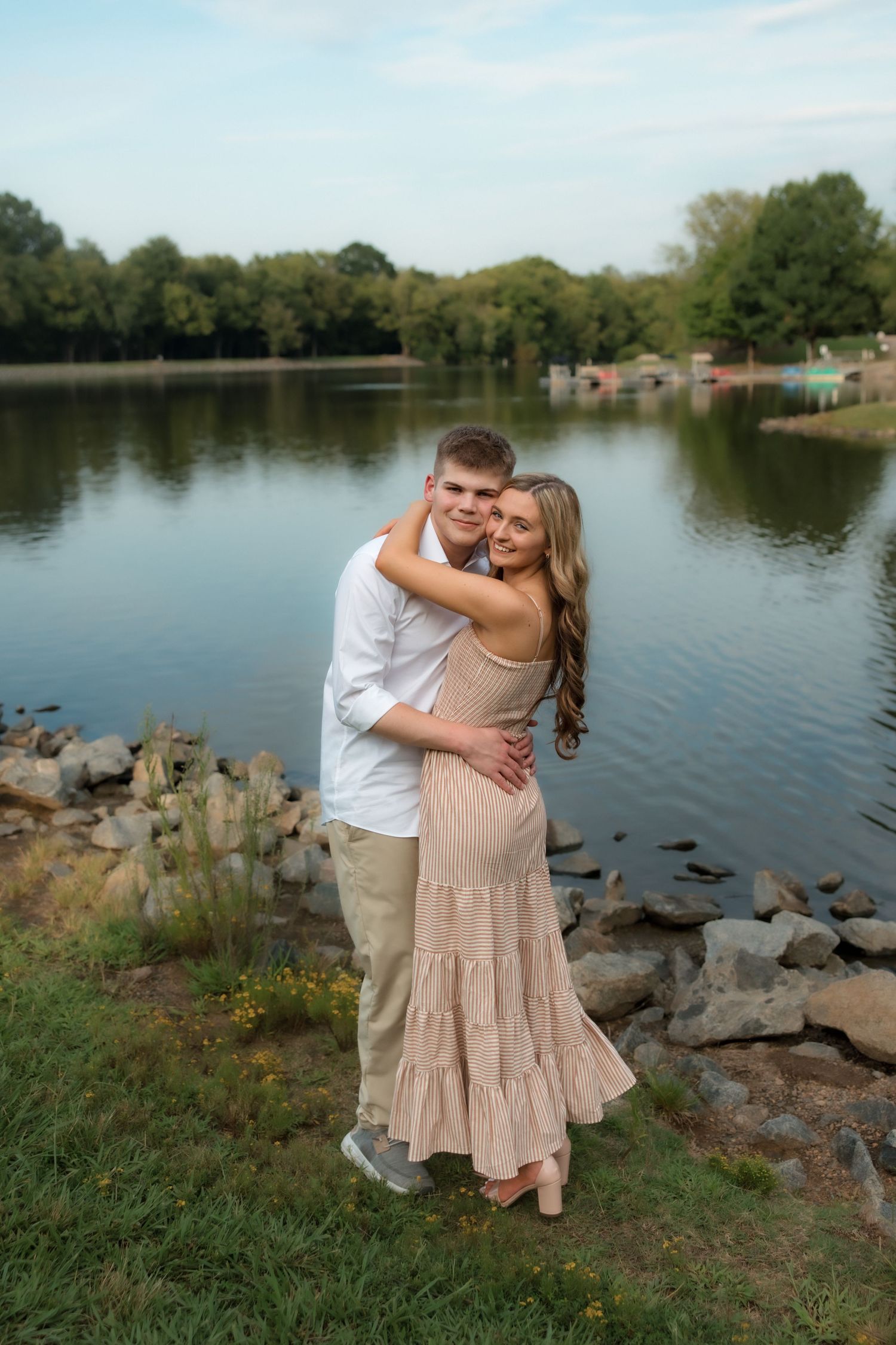 A couple embraces lovingly by a serene lakeside, surrounded by rocks and lush green trees.
