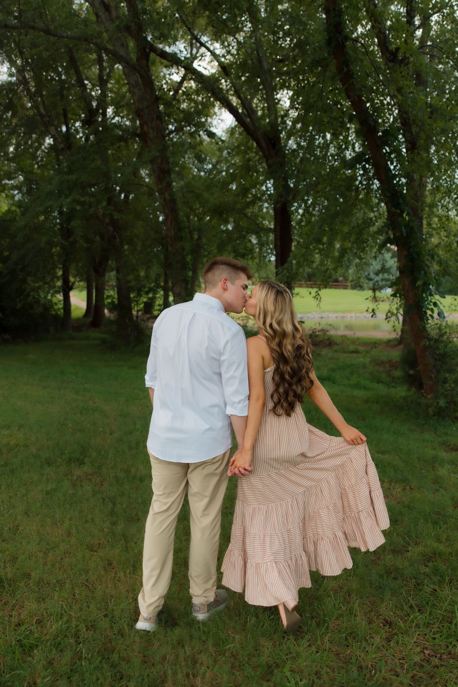 A couple sharing a kiss while holding hands in a lush green park, woman wearing a flowing blush maxi dress.