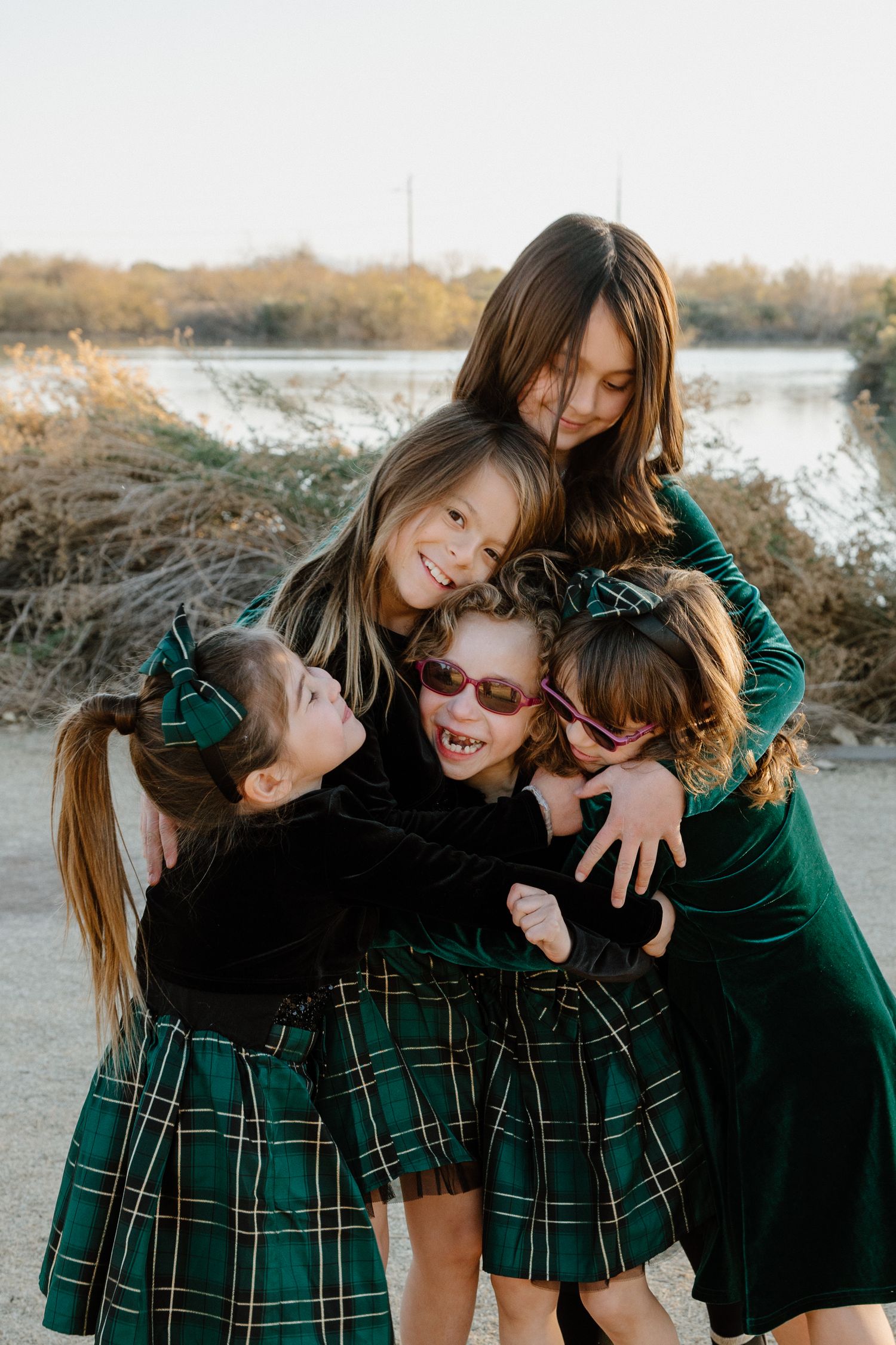 Five girls in matching green plaid dresses share a joyful group hug outdoors near a lake.