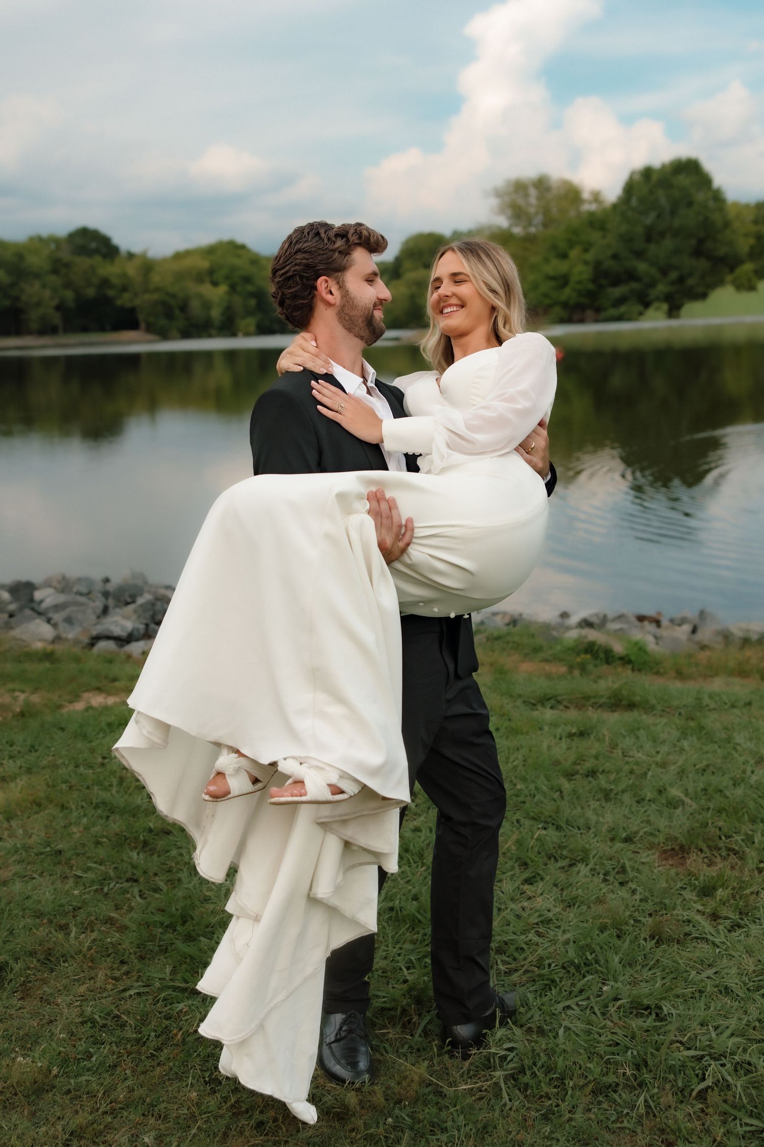 Smiling groom lifts bride in white dress by a serene lake surrounded by green trees.