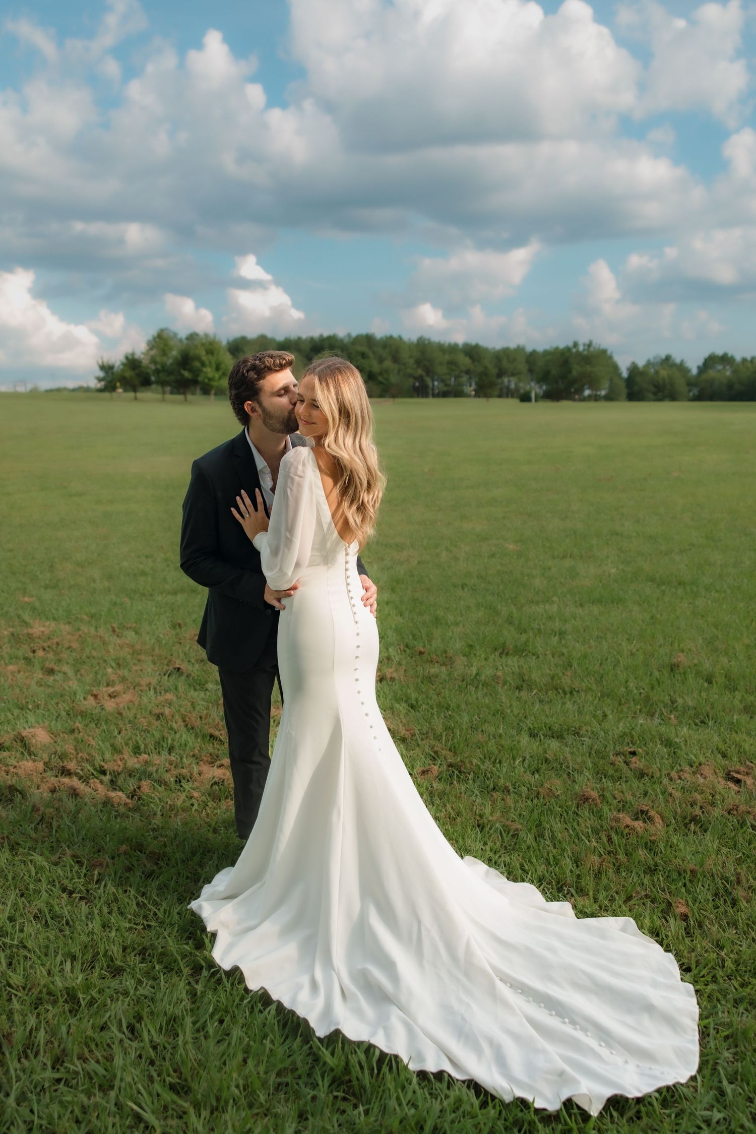 A bride and groom share a kiss in a lush green field, the bride wearing an elegant white mermaid gown with a long train.