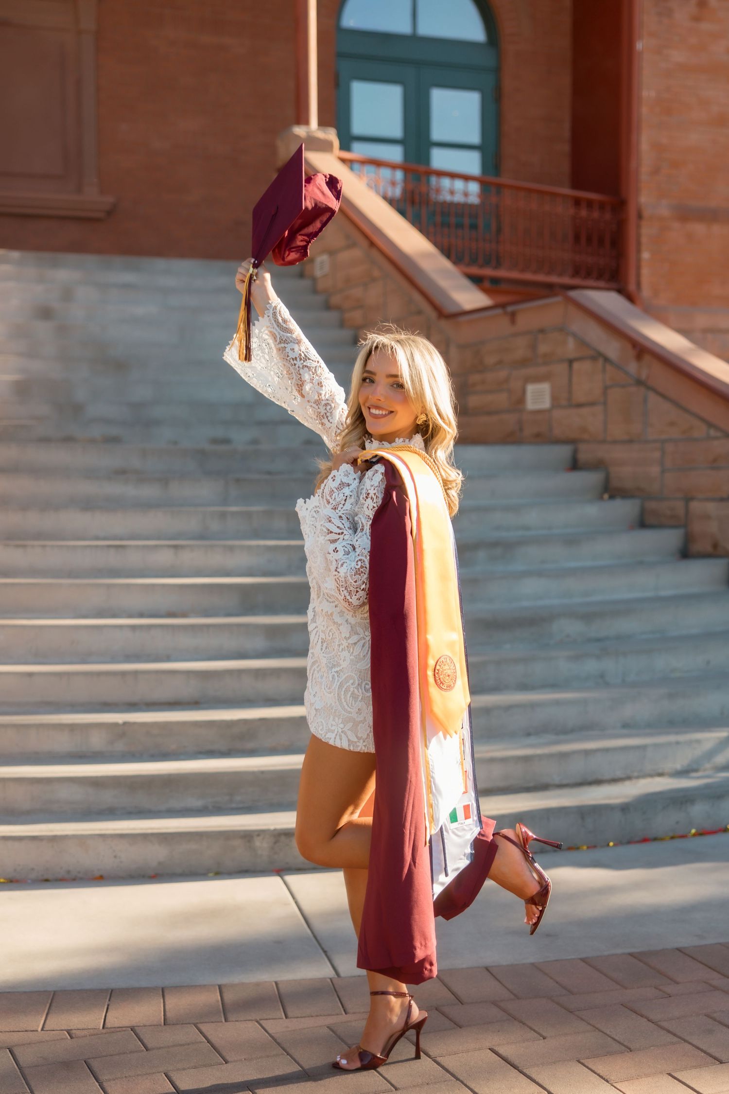 Smiling blonde graduate raises maroon cap on steps wearing white lace dress with gold and maroon stole.