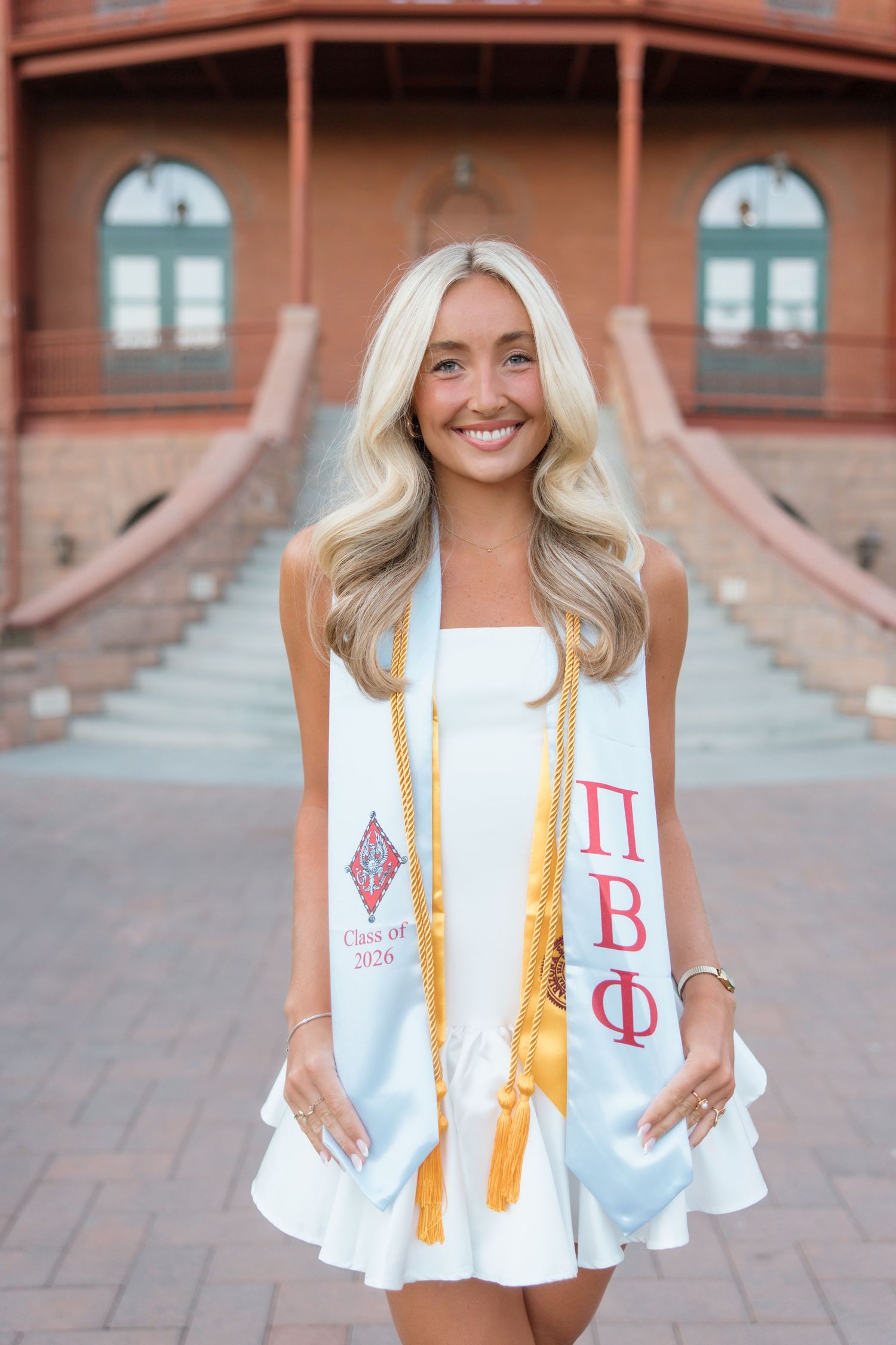 Smiling college graduate in white dress wearing Pi Beta Phi sash and gold honor cords poses outdoors at campus.